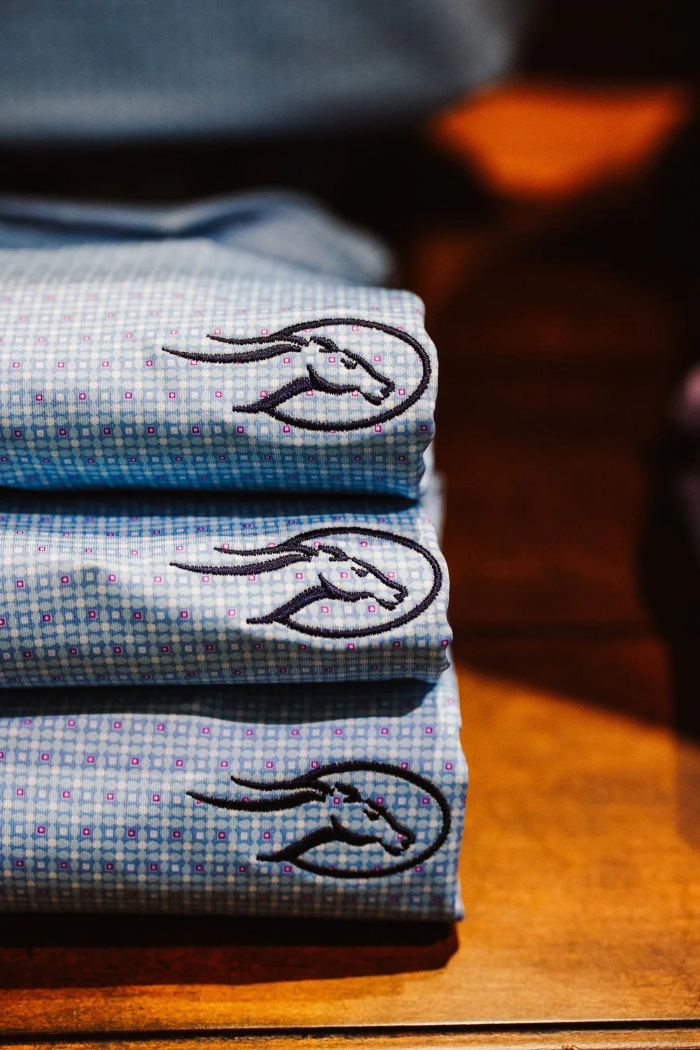 Stacked folded blue cloth face masks with a black embroidered horse logo, placed on a wooden surface.