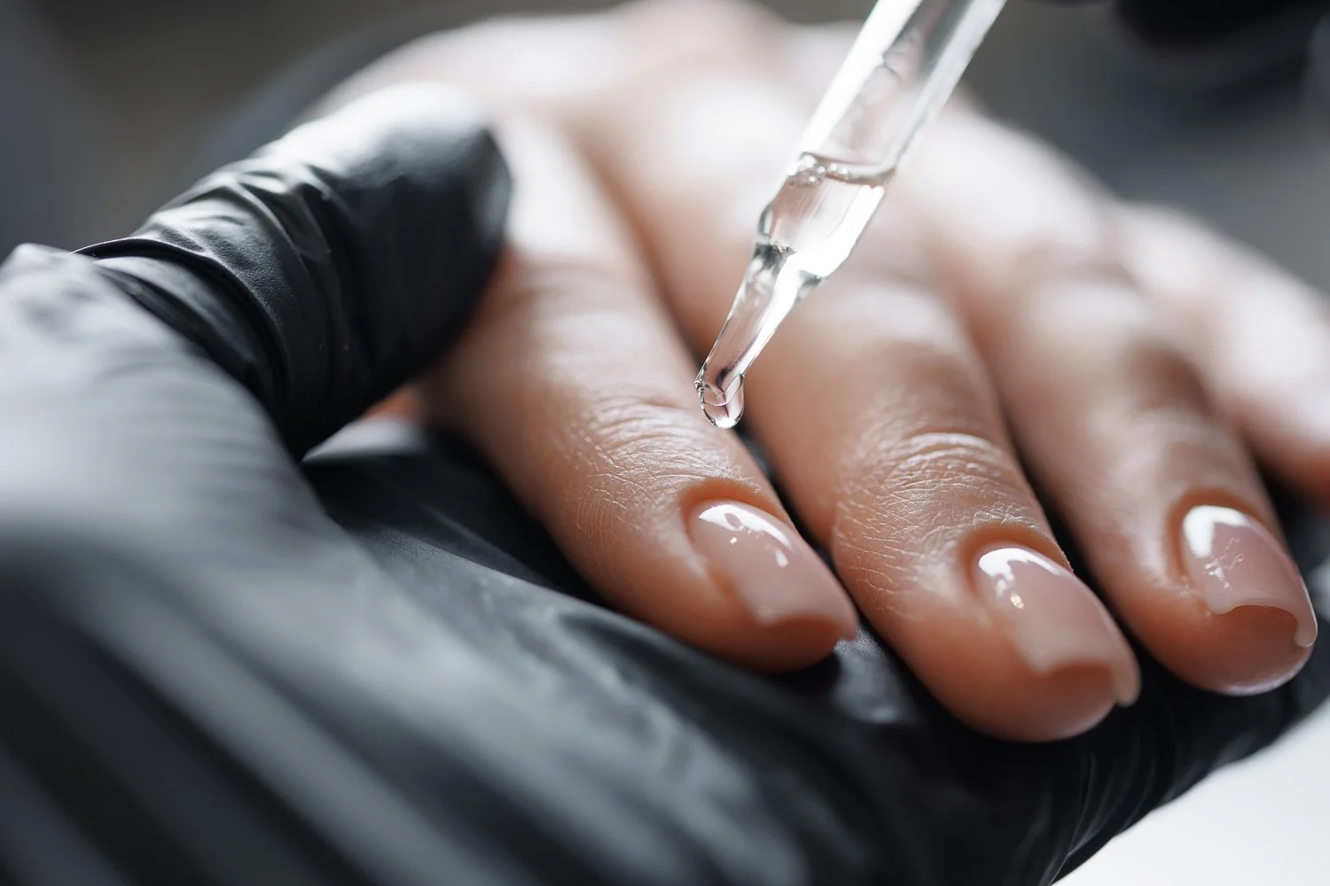 A person receiving a manicure, with a close-up of a hand with neatly polished nails and a dropper applying a clear liquid to the nails.