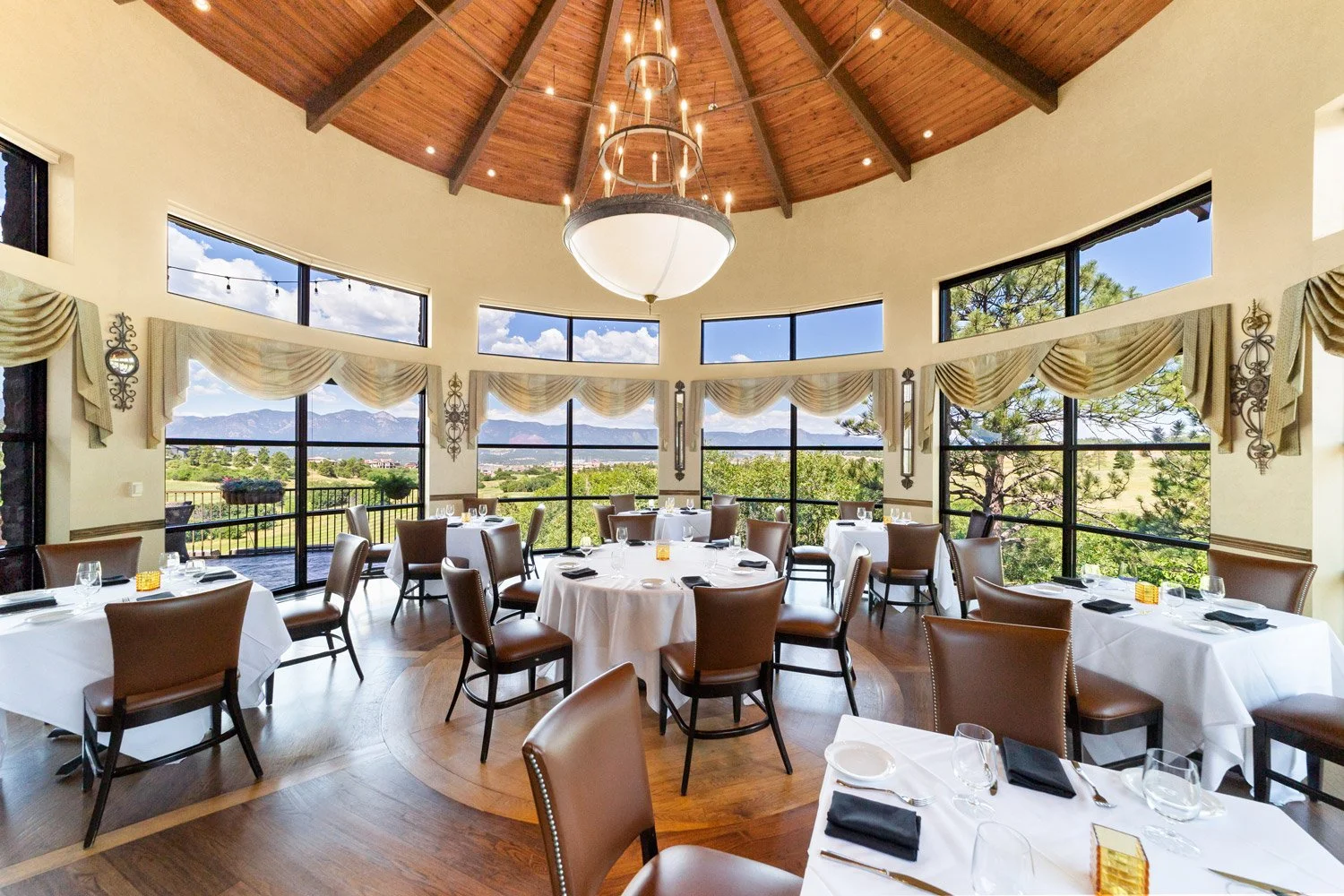 Elegant dining room, at The Steakhouse, with large windows displaying a scenic mountain view, round tables with white tablecloths, brown chairs, and decorative curtains.