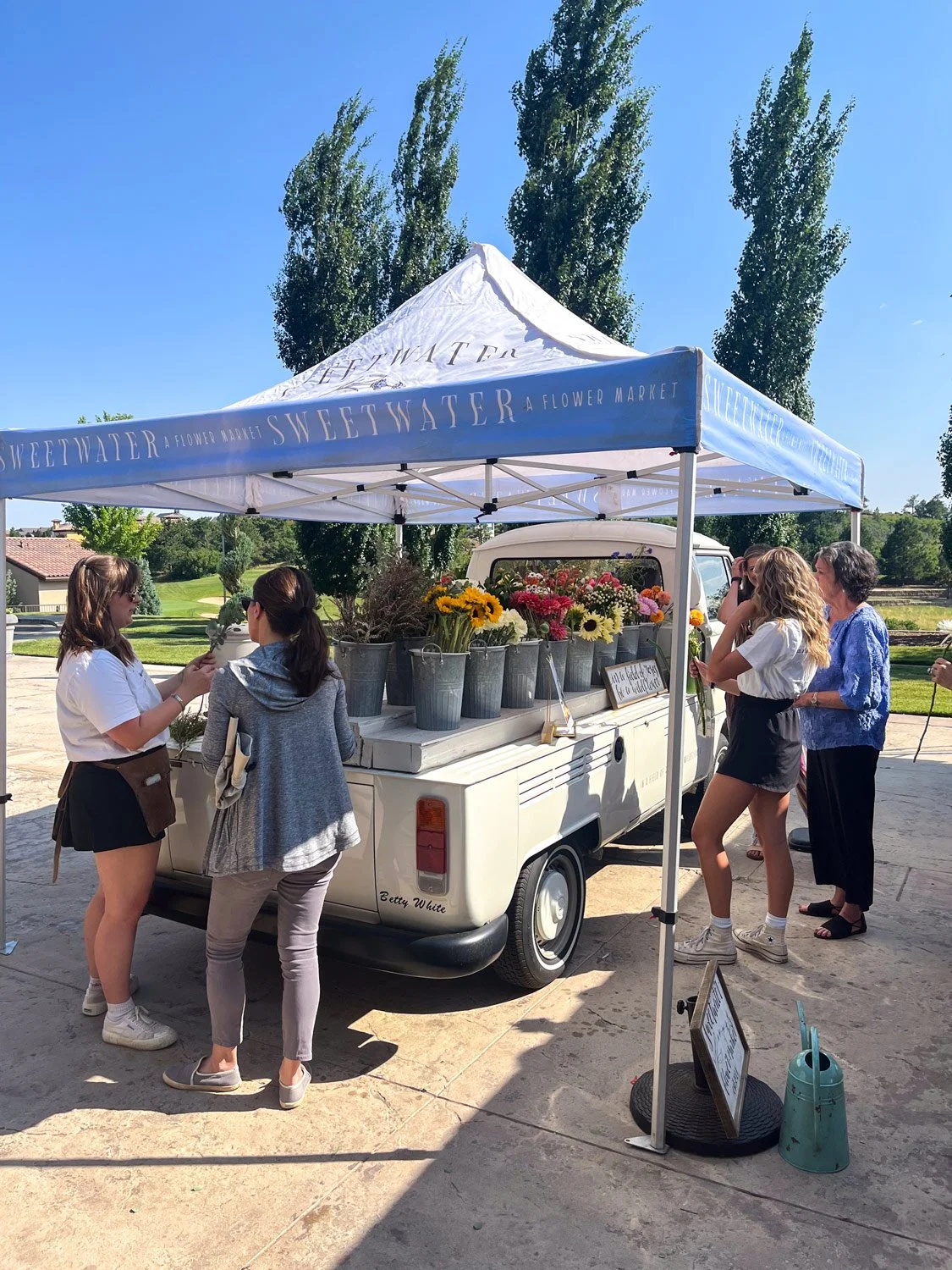 People shopping for flowers at a flower stand under a blue and white Sweetwater Flower Market canopy on a sunny day.