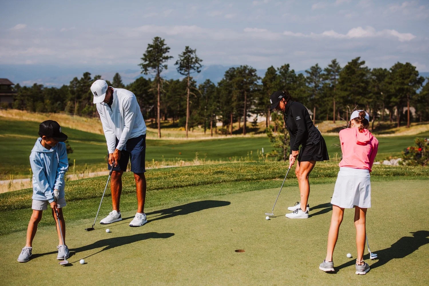 A family practicing golf on the Flying Horse North putting green under a partly cloudy sky, with trees and hills in the background.