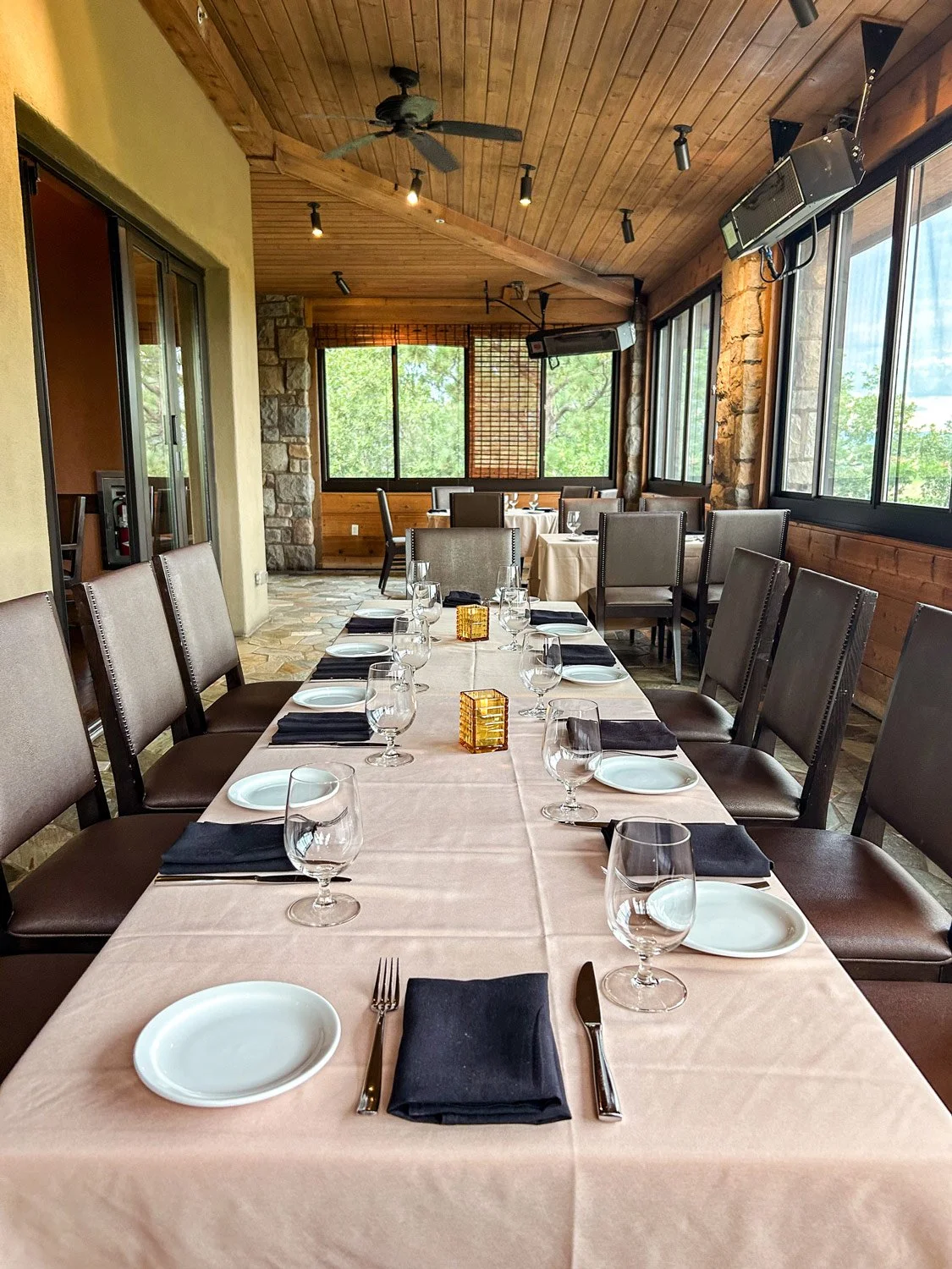 A restaurant dining room set for a special occasion, with a long table covered in a beige tablecloth, black napkins, white plates, wine glasses, and silverware. The room features large windows with natural light, a wooden ceiling with ceiling fans, a