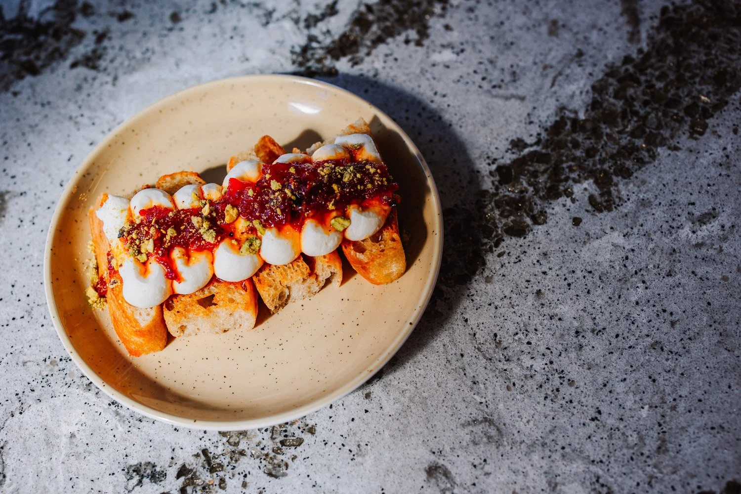 A plate of sliced toasted bread topped with dollops of cream, red sauce, and sprinkled pistachios, served on a speckled gray surface.
