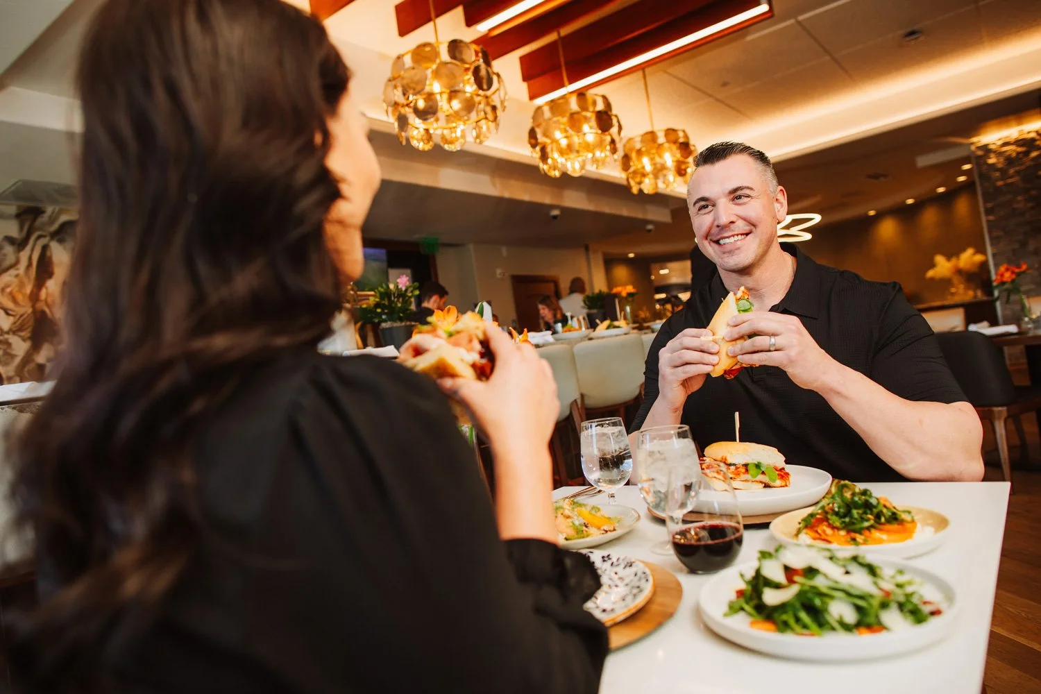 A man and woman enjoying a meal together at a restaurant, smiling and holding sandwiches, with various dishes and drinks on the table.