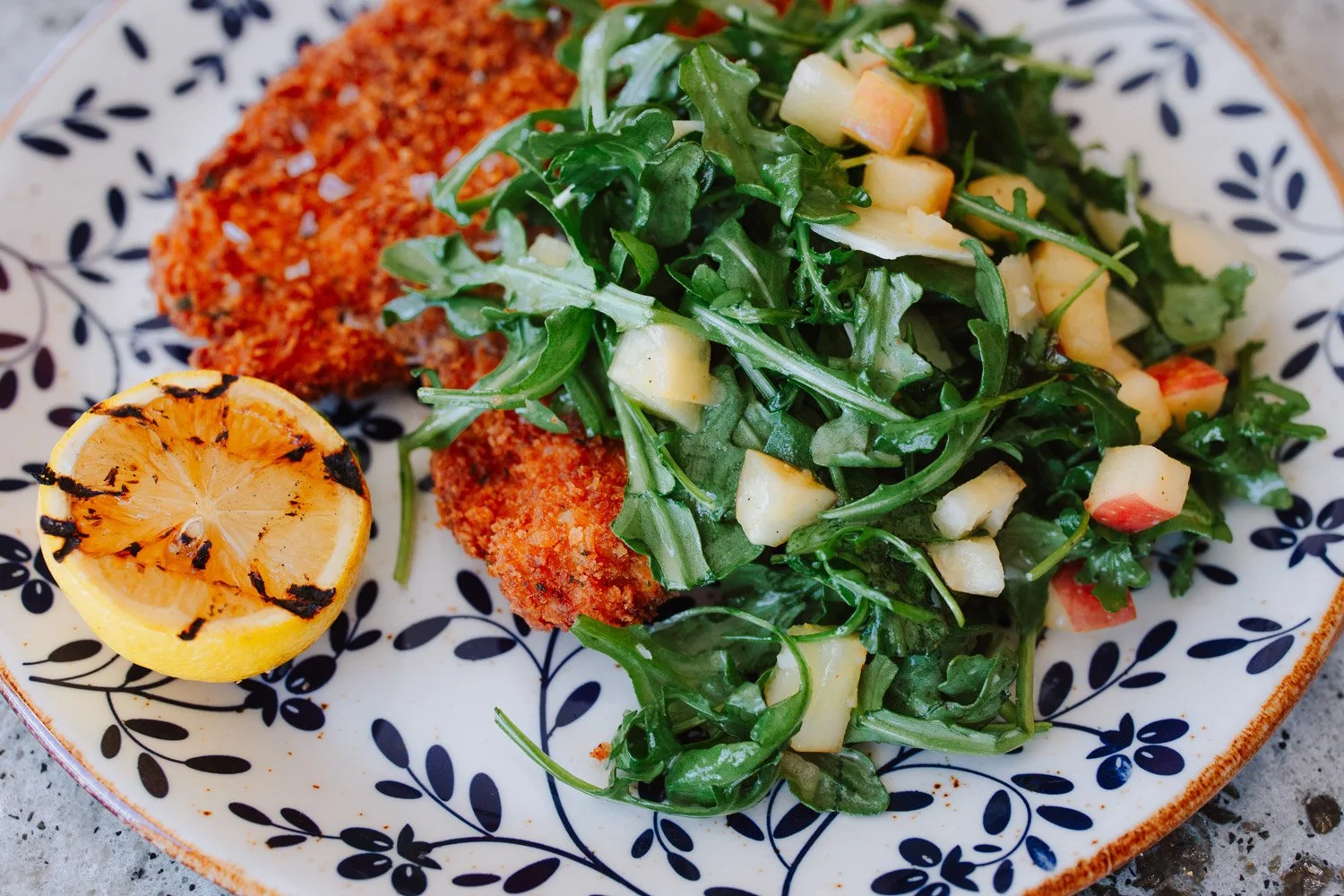 Plate with grilled lemon, arugula salad with chopped apples and tomatoes, and breaded chicken cutlets.