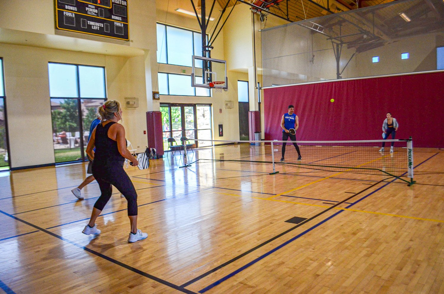 People playing pickleball on an indoor court with a basketball hoop and scoreboard in the background.