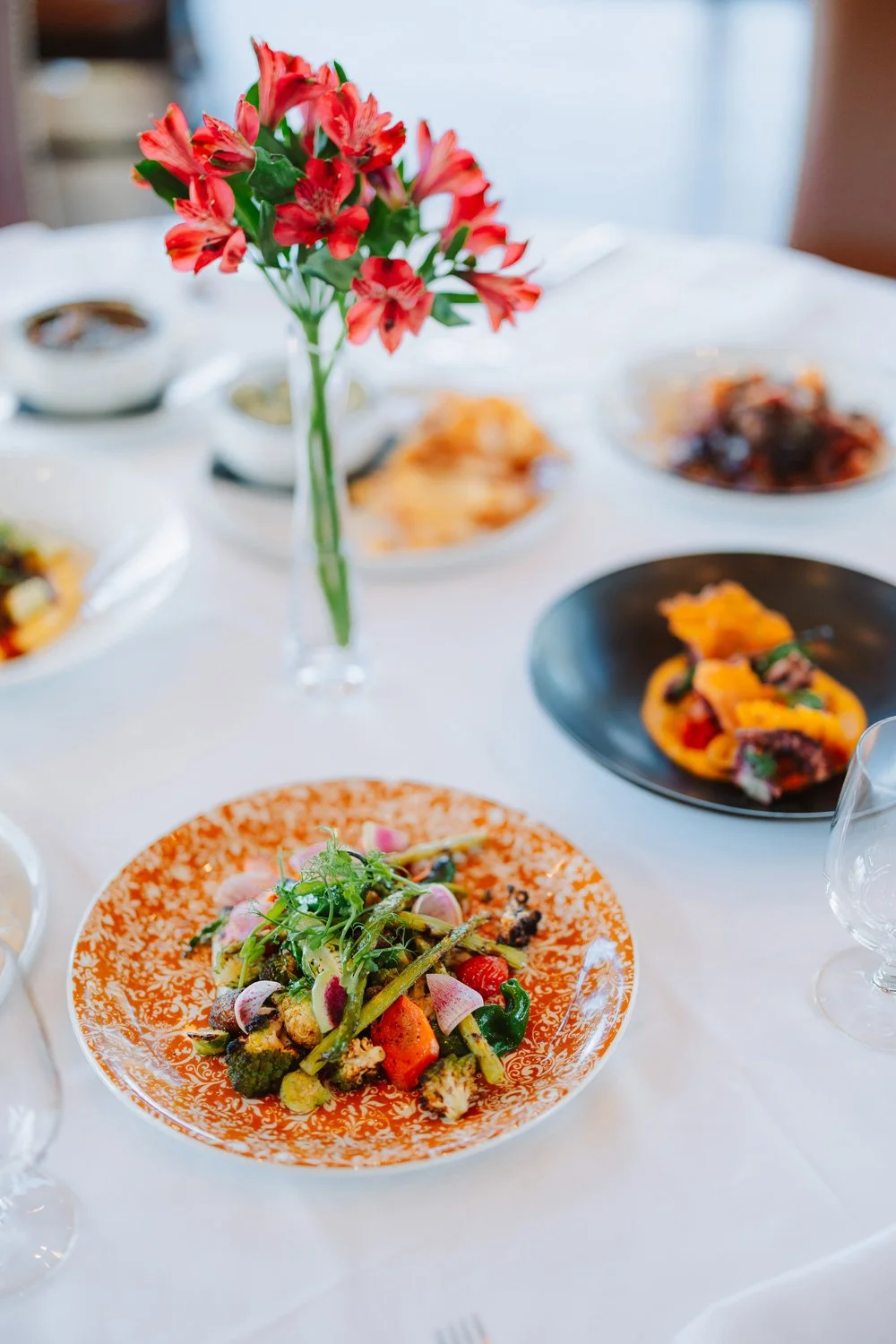 A table set with various plates of gourmet food and a glass vase with red flowers.