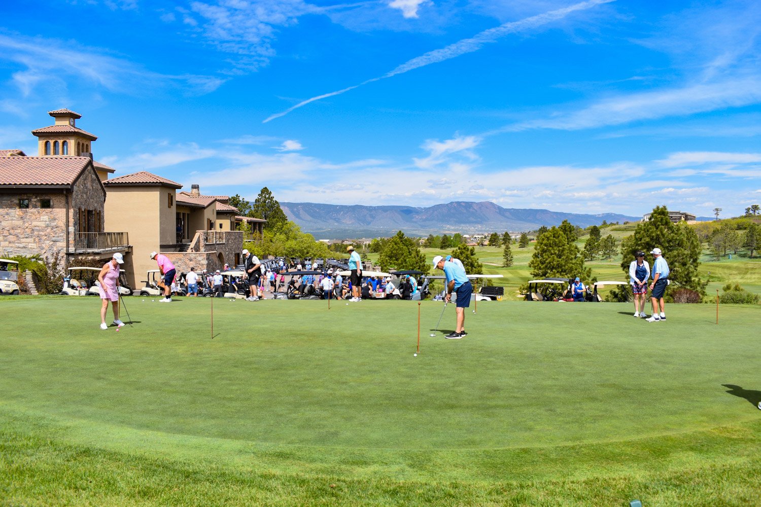 Golfers practicing putting on a course with a clubhouse and parking lot in the background under a blue sky with clouds and mountains.
