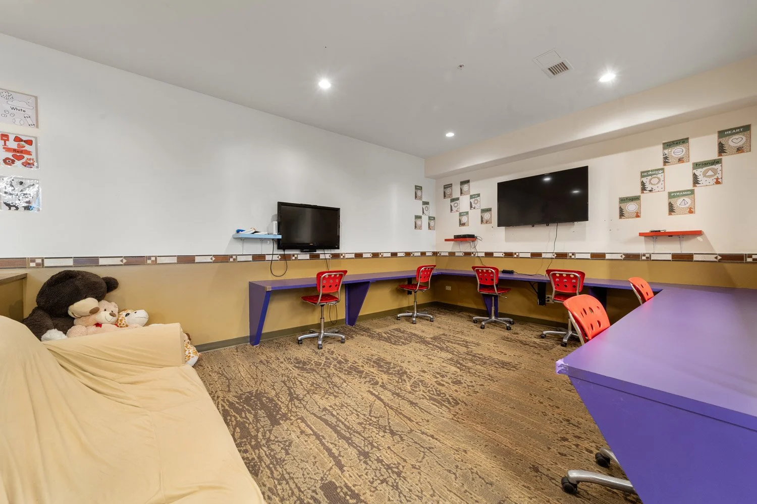 Classroom with striped beige and brown walls, red chairs, blue desks, two TV screens on wall, colorful alphabet posters, and plush teddy bears on beige couch