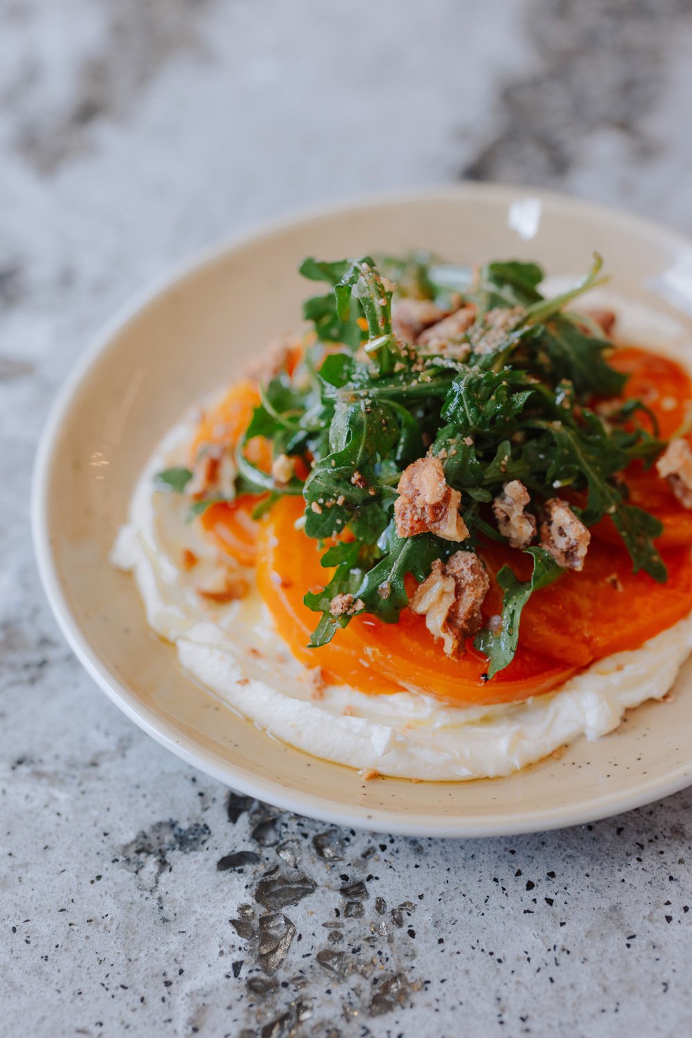 A plate of a fried egg, topped with arugula, crushed nuts, and a dollop of a creamy white sauce, served on a speckled gray surface.