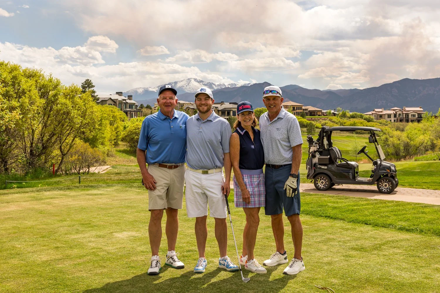 Group of four people smiling on a golf course with a golf cart nearby and mountains in the background.