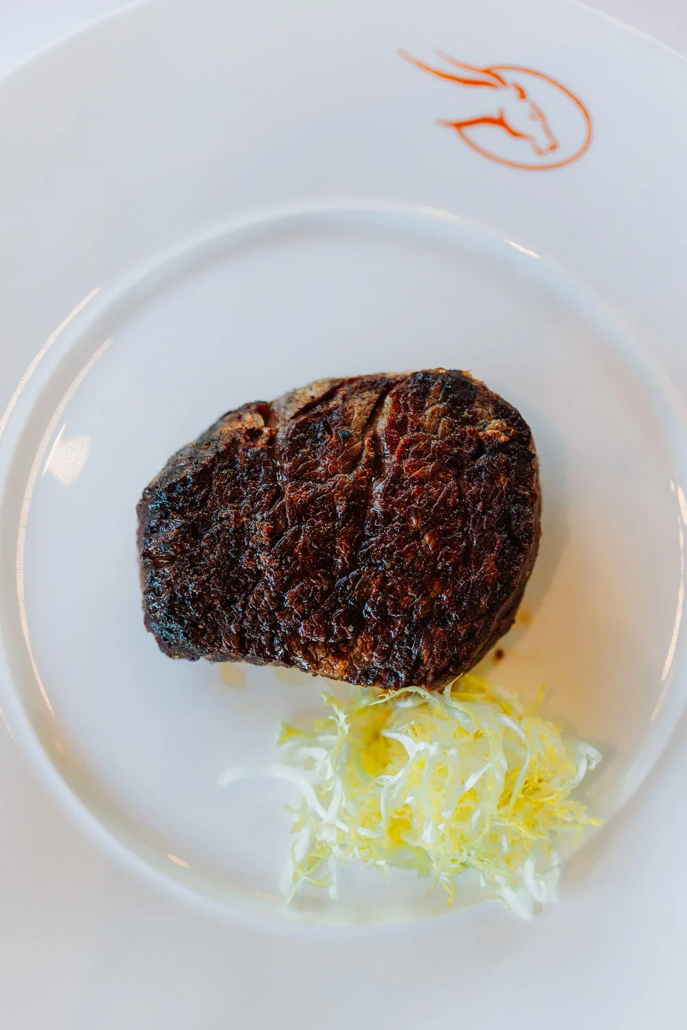 A steak on a white plate with a logo, accompanied by a small portion of shredded lettuce.
