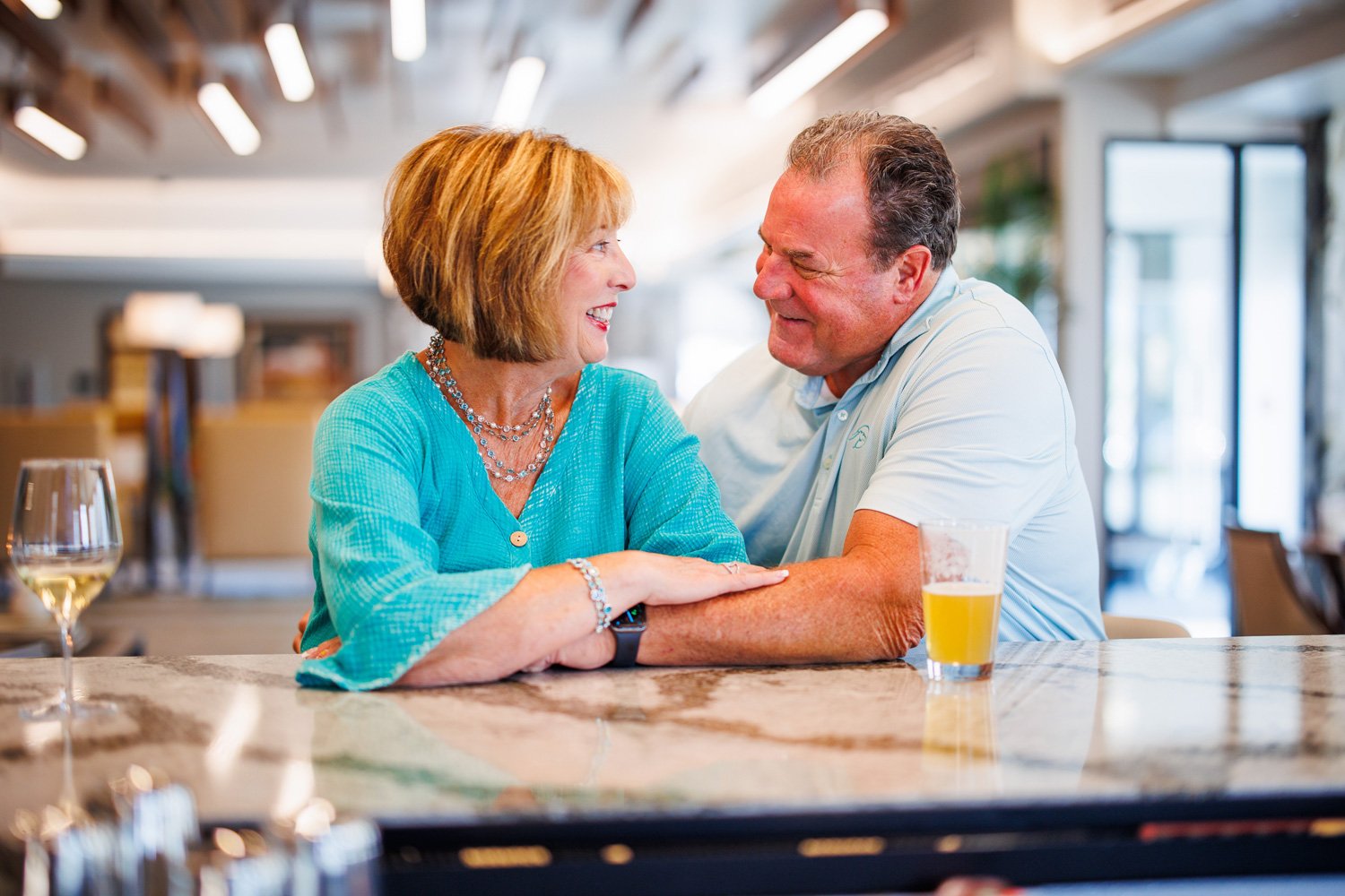 A smiling elderly woman and a middle-aged man sharing a joyful moment at a table in a bright, modern restaurant or cafe. The woman has reddish hair and wears a turquoise blouse and jewelry. The man has short dark hair and wears a light blue polo shir