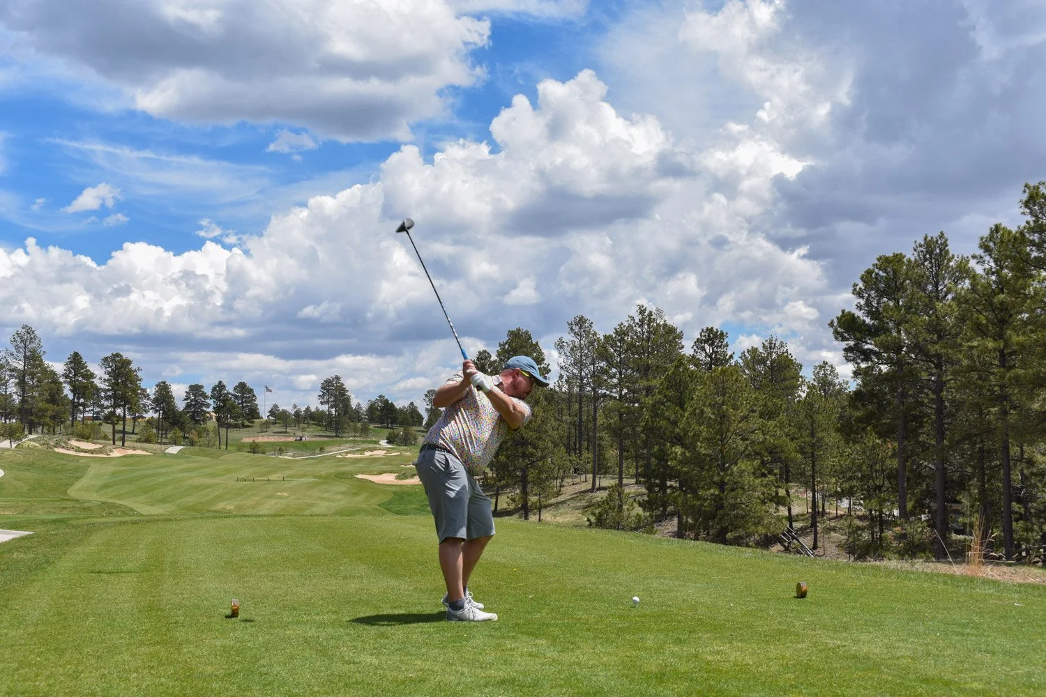 A man playing golf on a sunny day at a golf course with trees and sand traps, in the background cloudy sky.