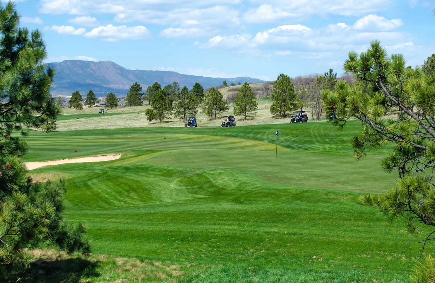 A golf course with green fairways, sand traps, and a flagstick, surrounded by trees and distant mountains under a blue sky with clouds.