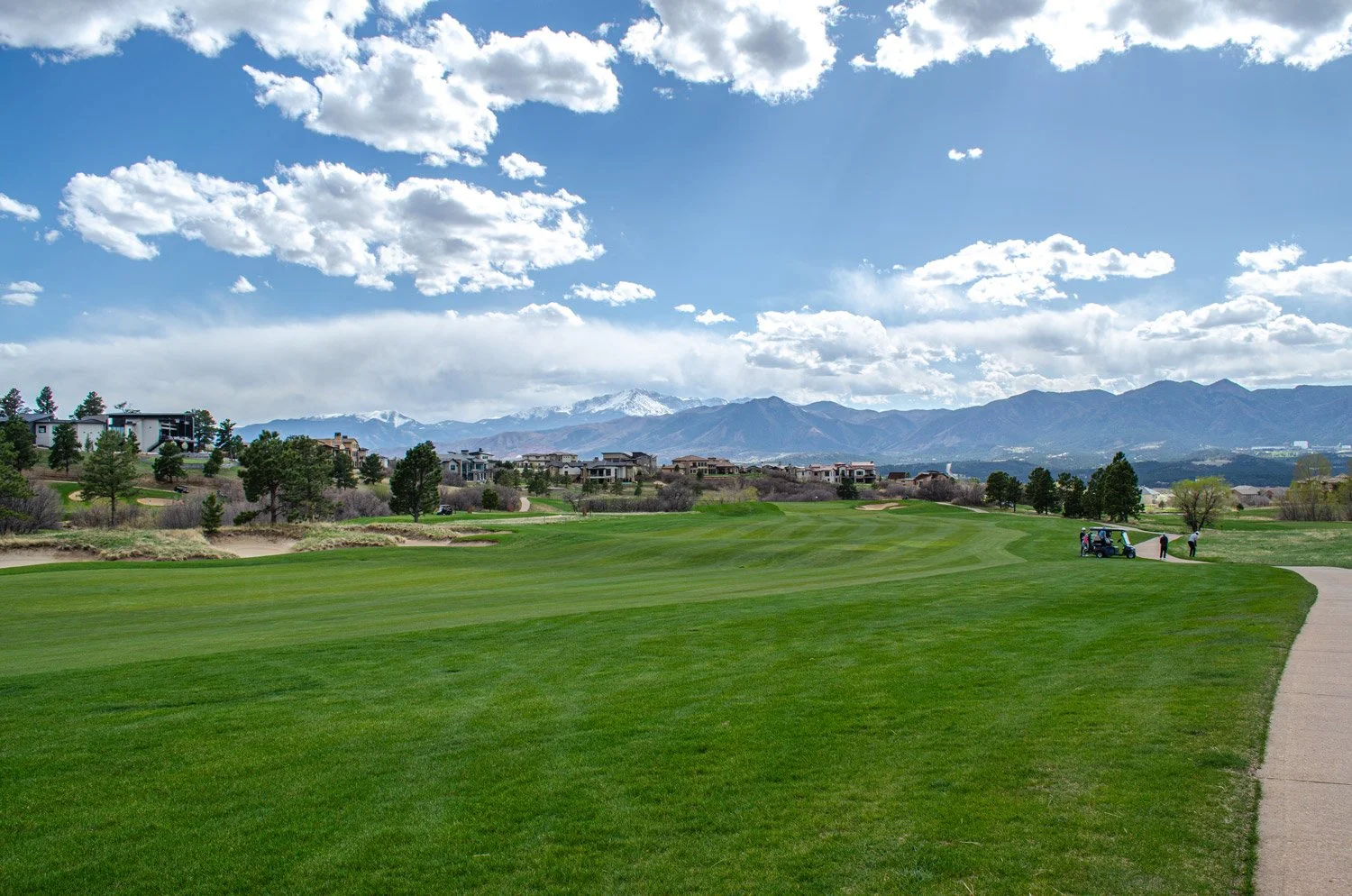 A golf course with green grass, a cart path on the right, people near a golf cart, trees, residential buildings, mountains in the background, and partly cloudy sky.