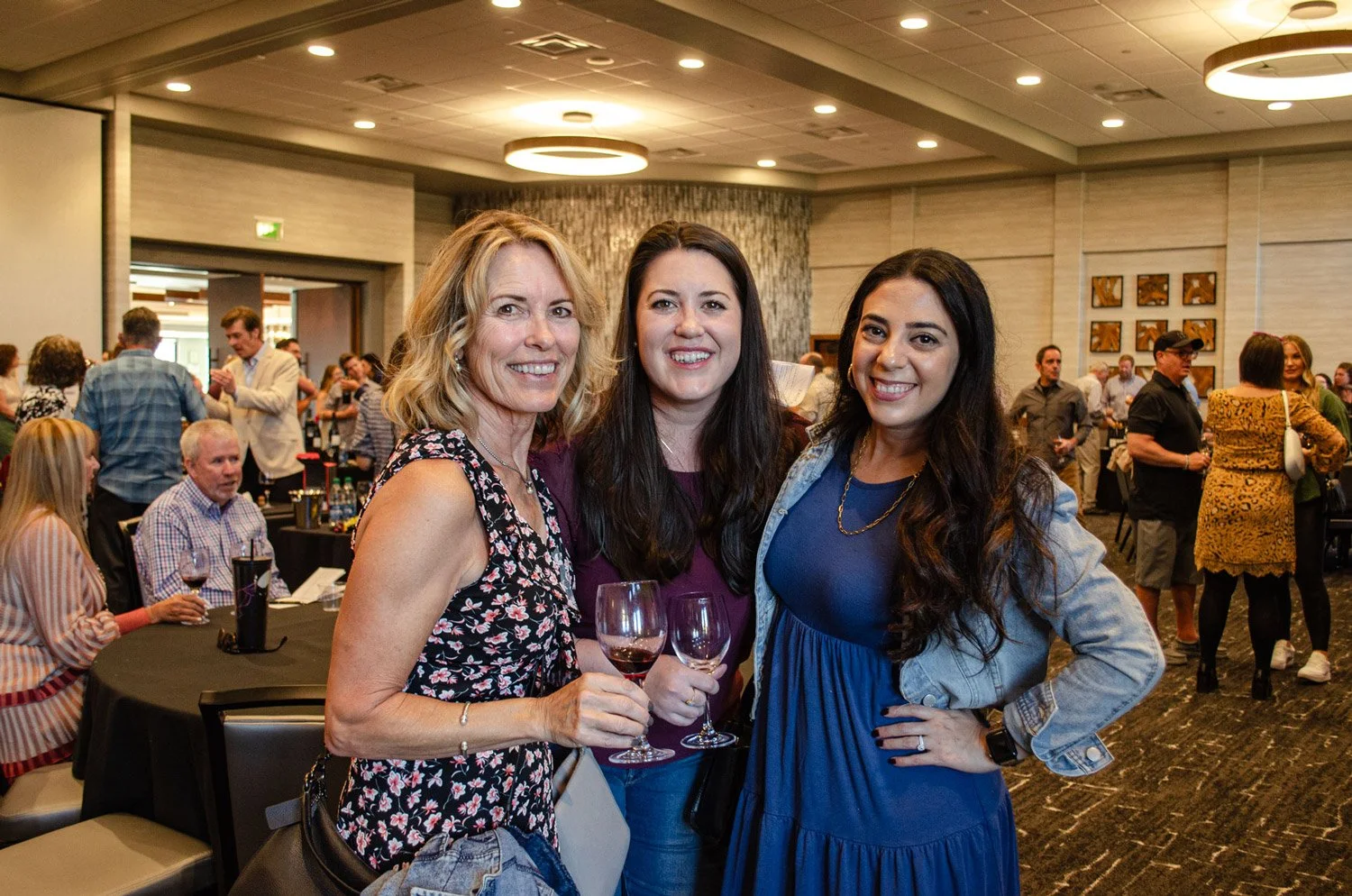 Three women smiling and holding glasses of red wine at a social event in a large room with many people and tables.