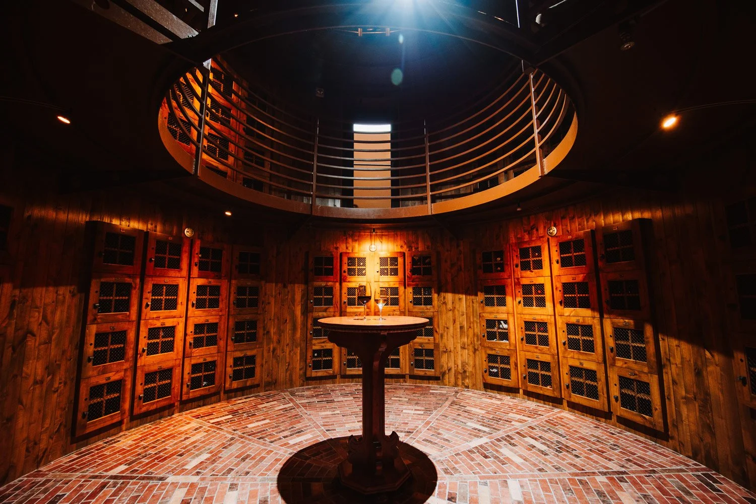 A wine cellar with wooden lockers lining the curved wall, brick flooring, a round table in the center with two wine glasses, a small wine bottle, and a glowing ceiling light.