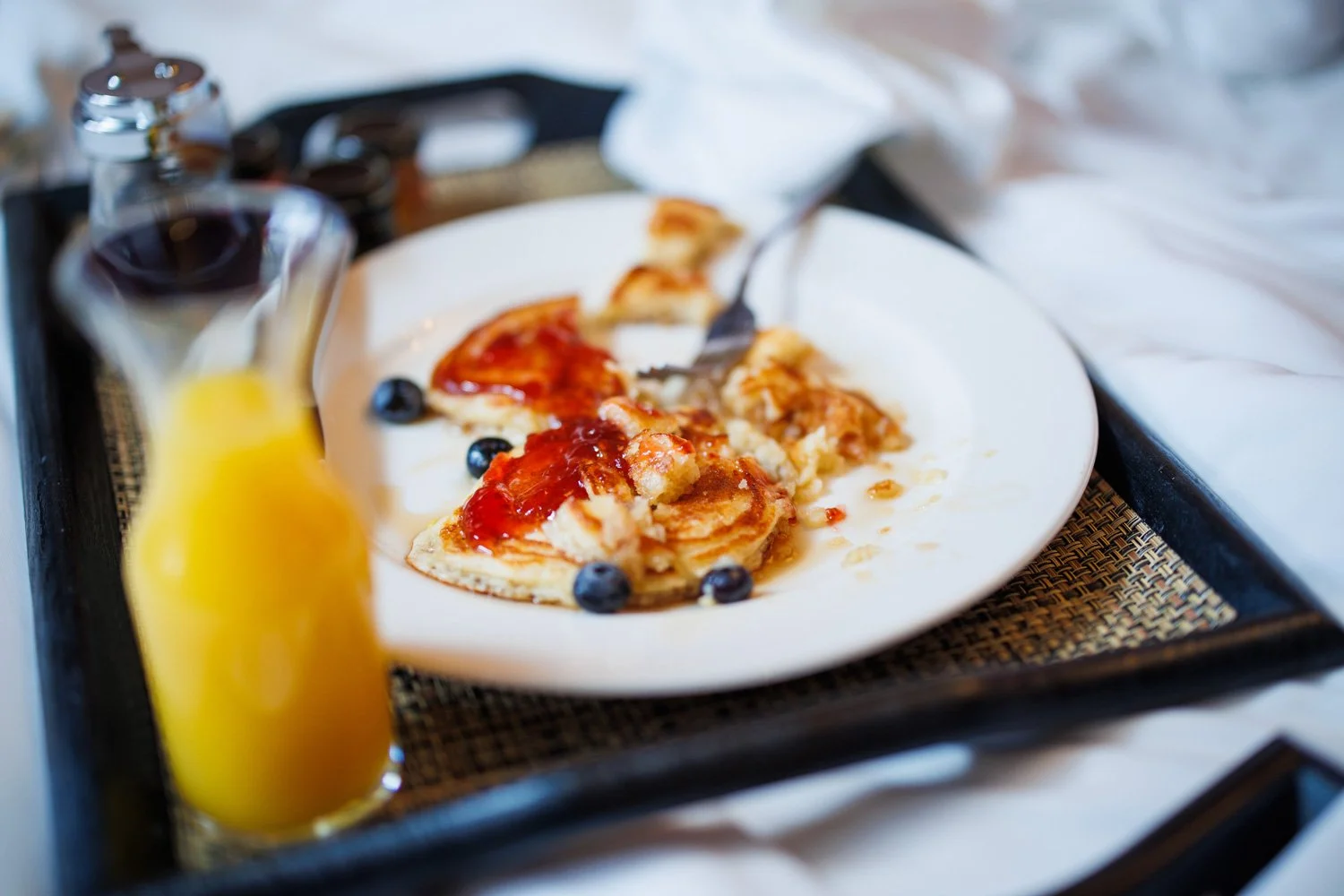 A tray with a white plate of leftover pancake with berry toppings, a small pitcher of orange juice, and a bottle of syrup on a woven placemat, with a blurred background.