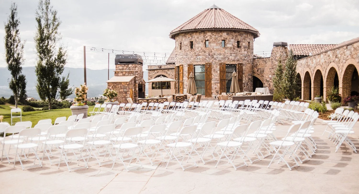 Outdoor wedding ceremony setup on the courtyard with rows of white chairs facing a stone building with a tower, in a landscaped area with trees and mountains in the background.