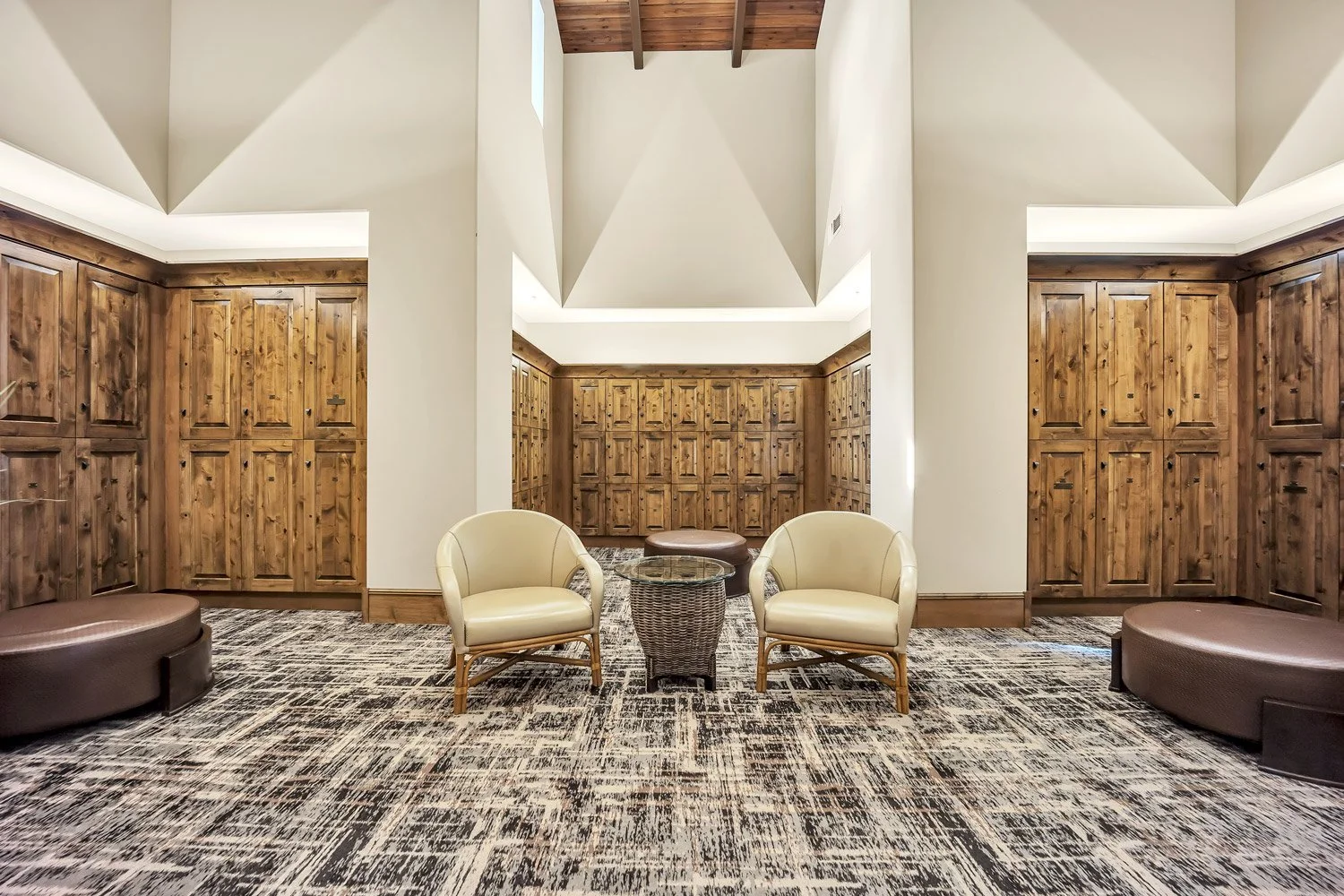 A cozy women's locker room lobby area with two beige armchairs, a glass-top table, and a woven side table between them, surrounded by wooden lockers and a patterned rug.