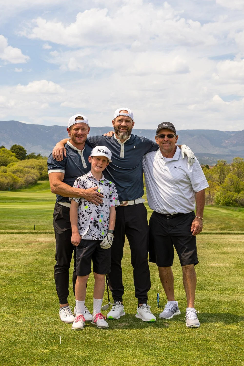 Four men and one young boy standing together on a golf course, smiling with mountains and partly cloudy sky in the background.