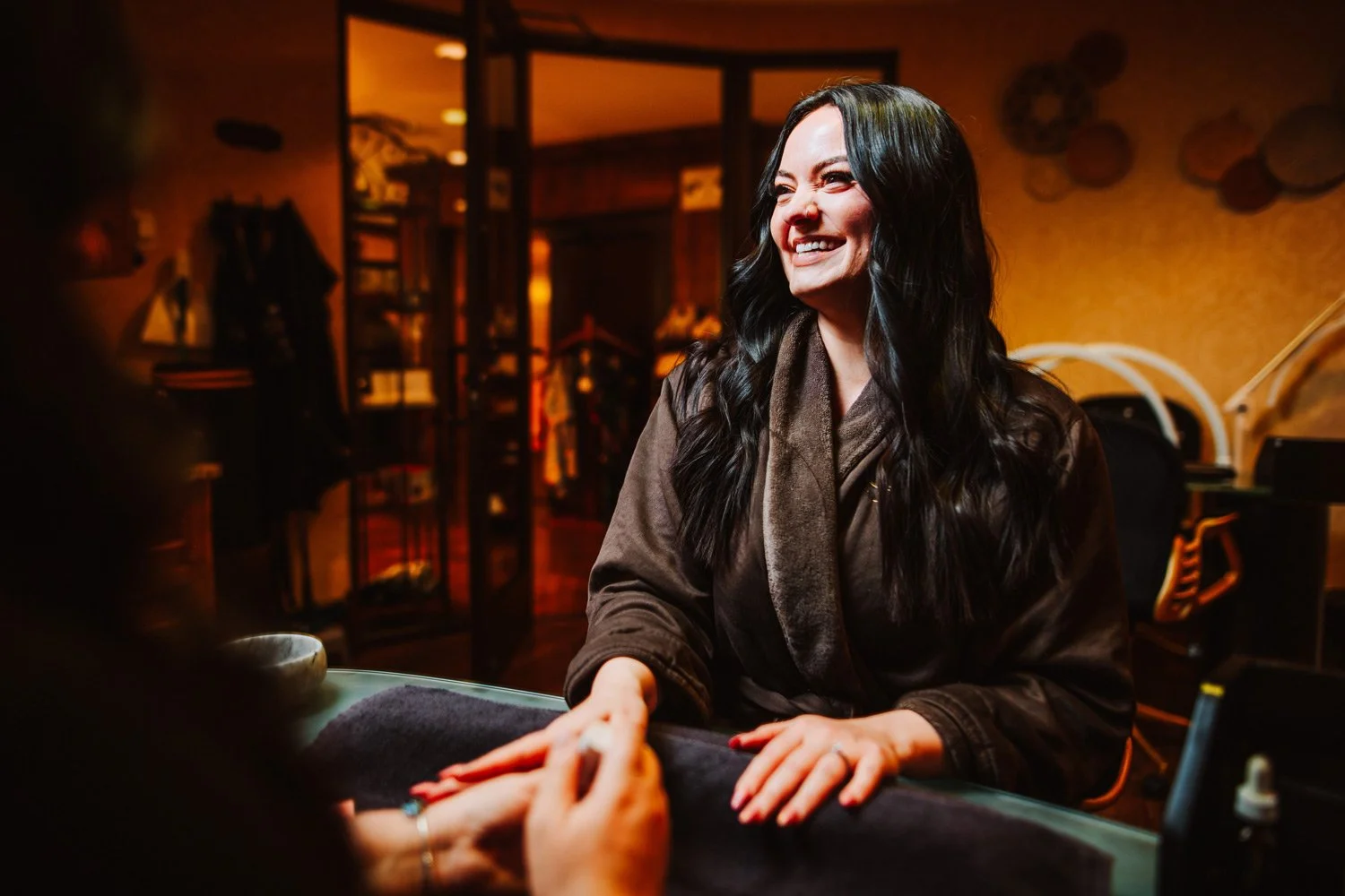 A woman with long dark hair smiling and sitting at a manicure table, receiving a manicure from a technician in a cozy, warmly lit spa treatment room.