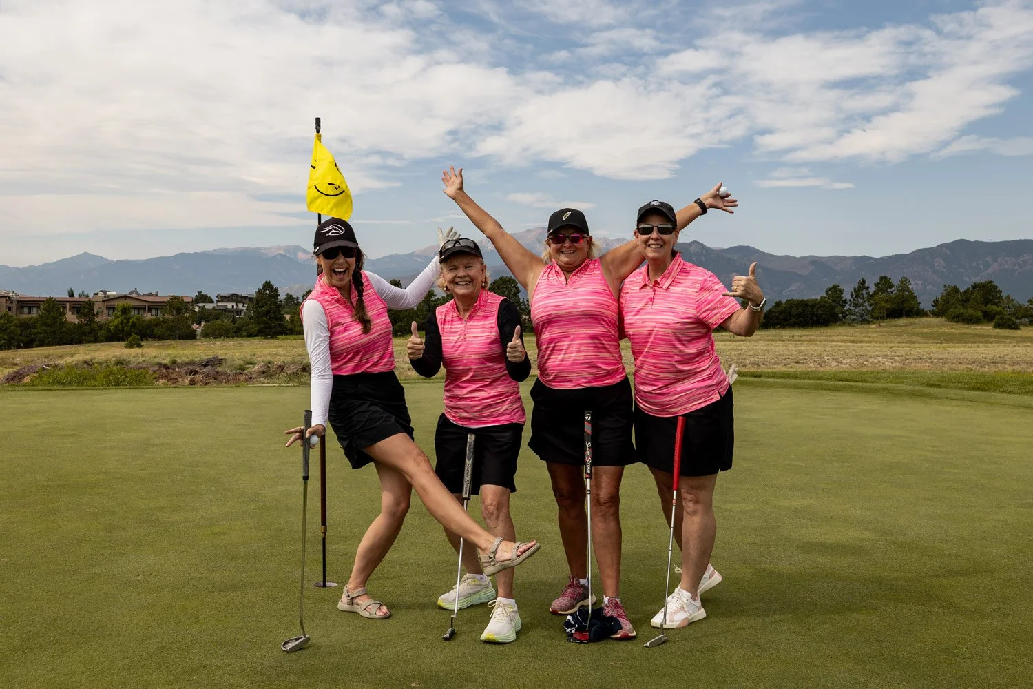 Four women in pink striped golf shirts and black shorts celebrating on the golf course, with mountains and a cloudy sky in the background.
