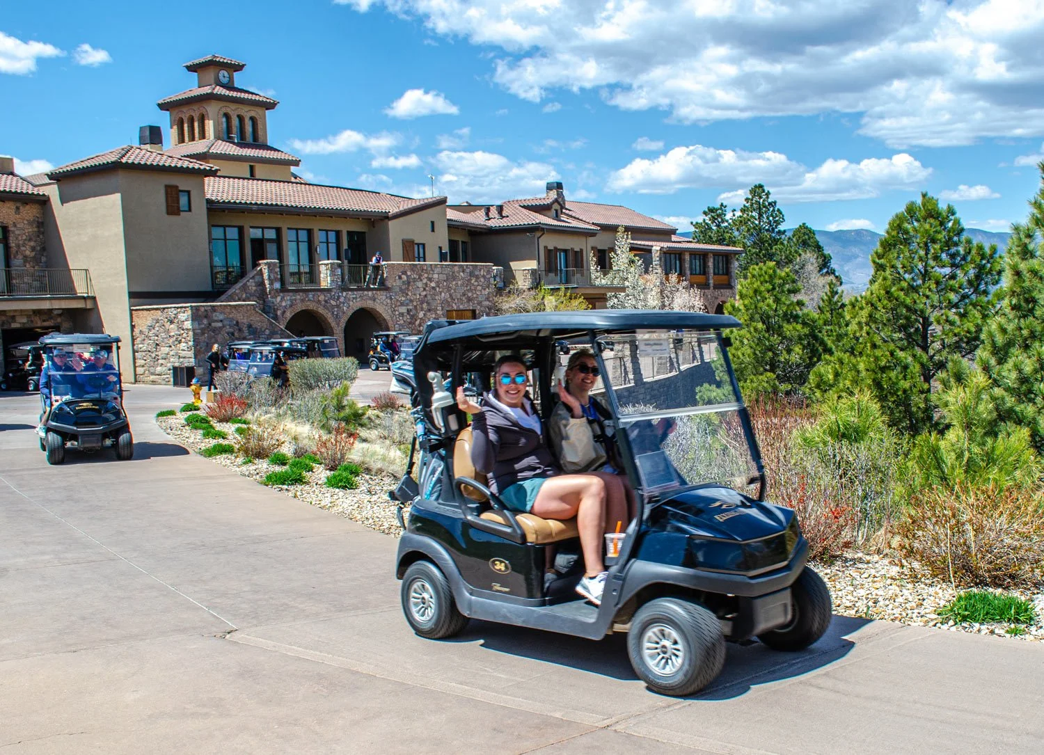 A group of people riding in a golf cart on a paved path outside a large building with a stone facade, surrounded by trees and shrubs, under a partly cloudy sky.