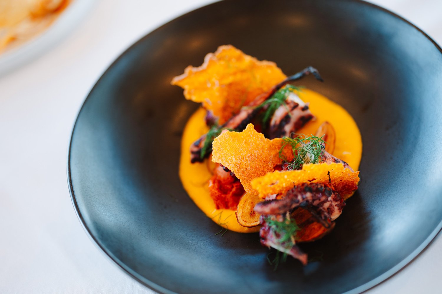 Gourmet seafood dish with grilled octopus, crispy potato chips, and a bright yellow sauce, garnished with fresh herbs on a black plate.