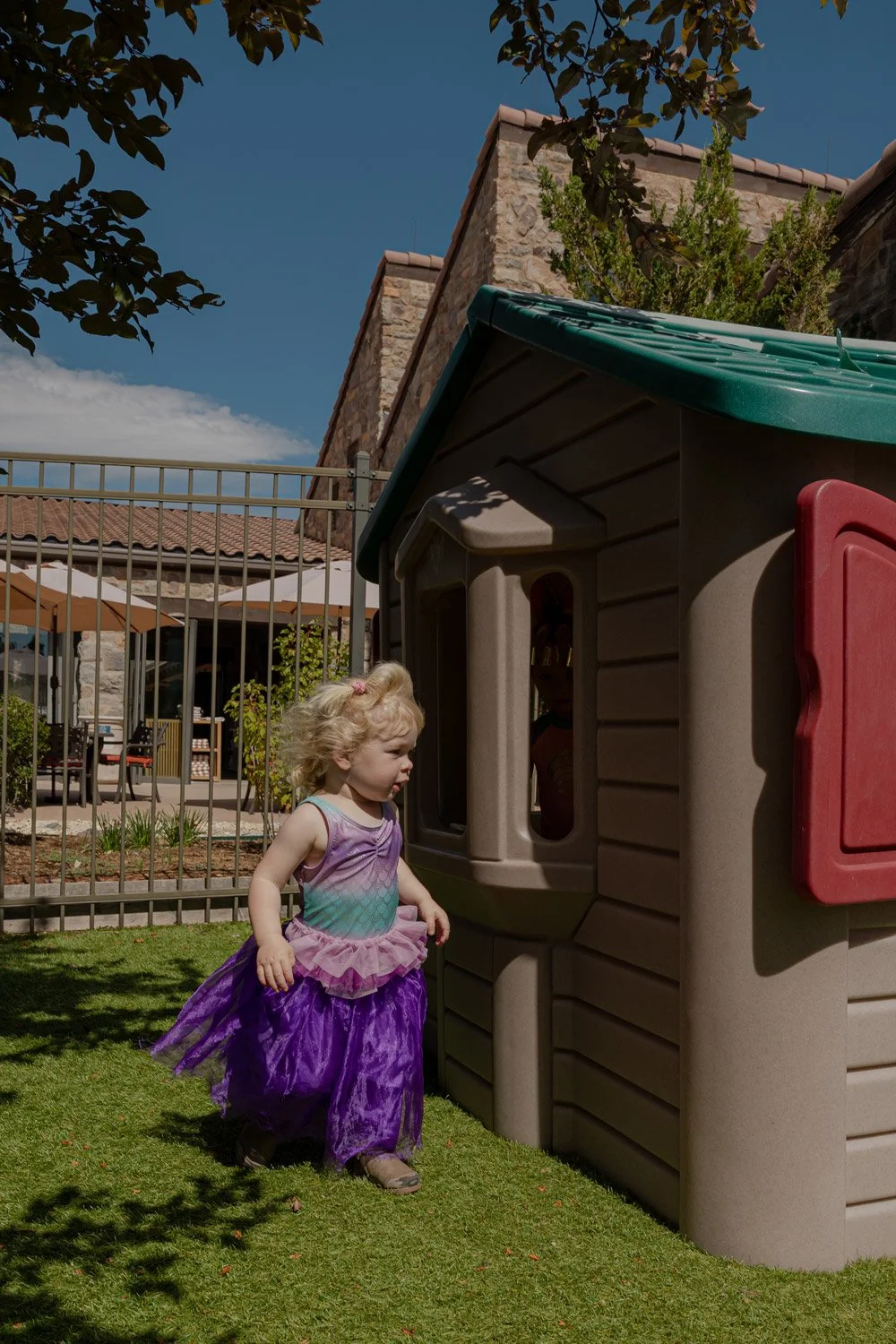 A young girl in a purple princess dress standing outdoors next to a playhouse on a sunny day.