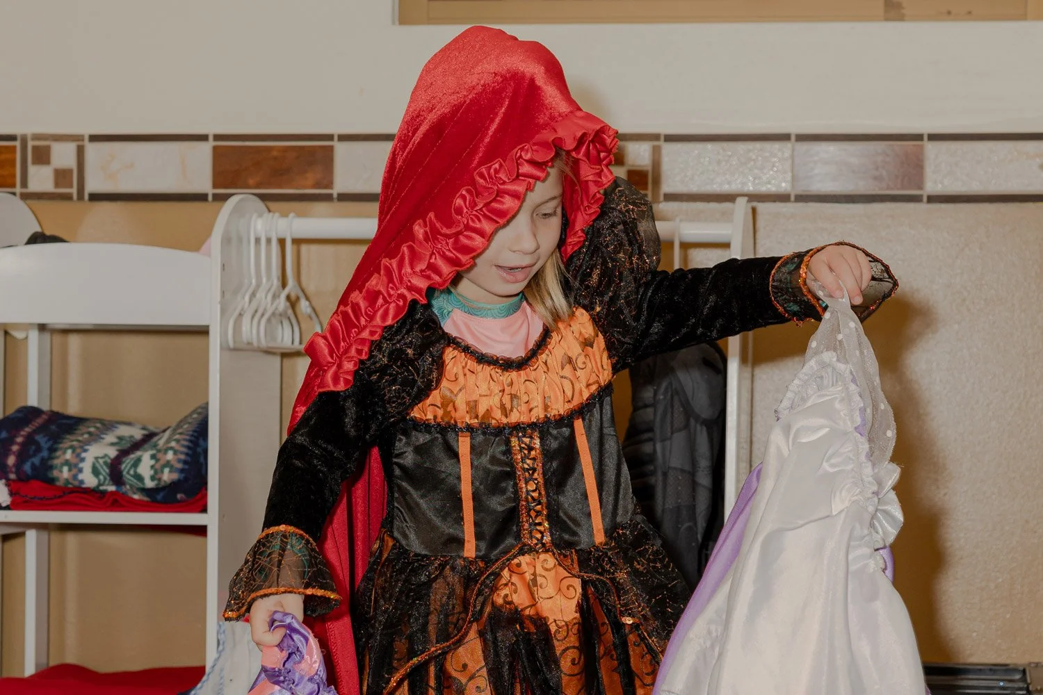 A young girl dressed as a witch, wearing a black and orange costume with a red hood, looks at a white dress with purple accents.