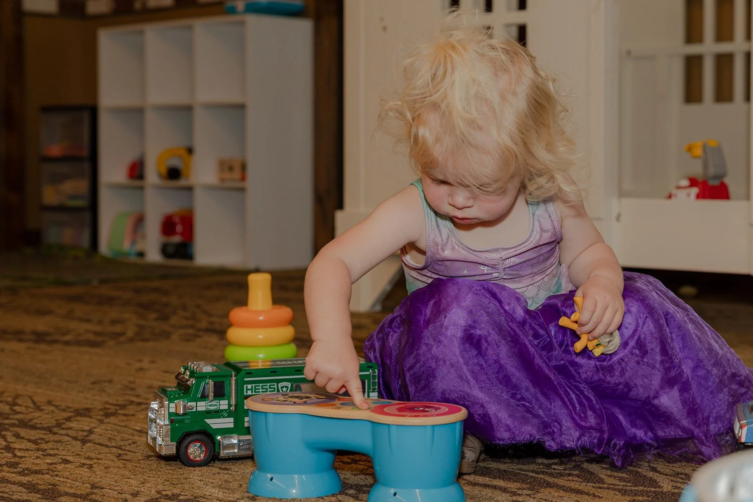 A young girl with blonde curly hair playing with toys on a carpeted floor in a playroom, including a green toy truck, a stacking ring toy, and a purple dress.