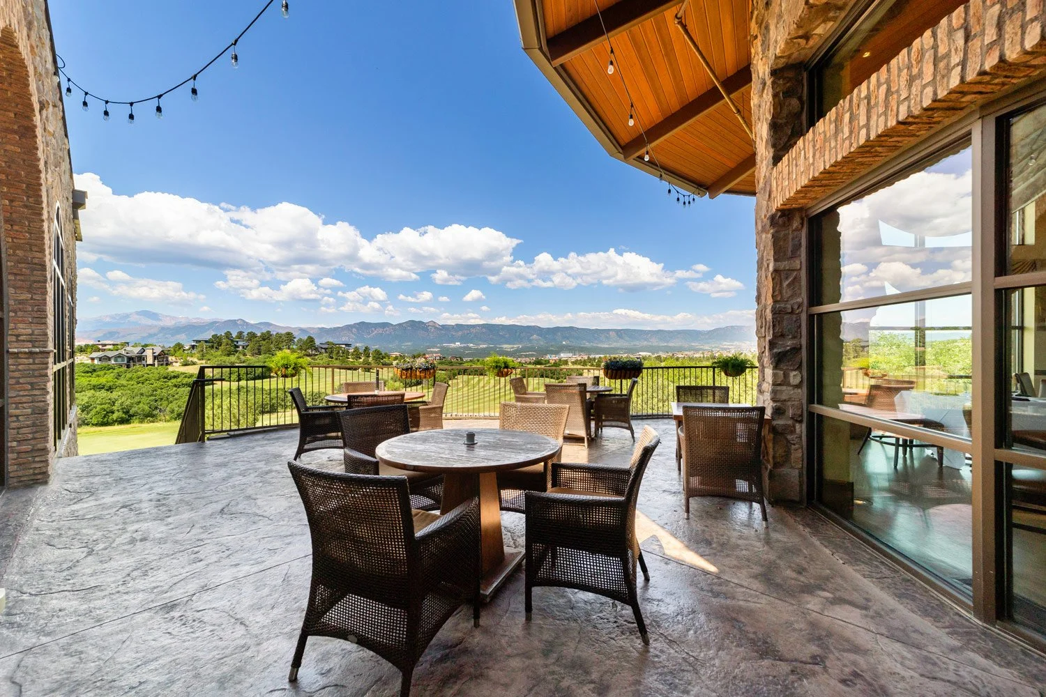 Outdoor patio with wicker chairs and round tables, overlooking a scenic landscape with mountains, green fields, and a bright blue sky with scattered clouds at The Steakhouse.