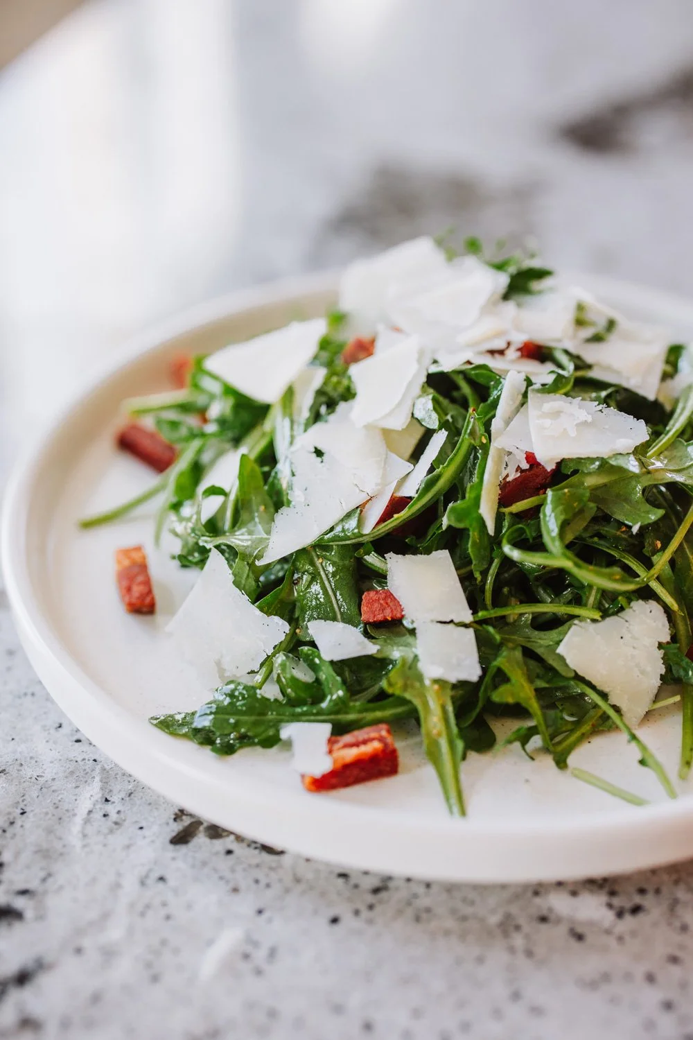 Fresh arugula salad topped with shaved Parmesan cheese and crumbled bacon on a white plate