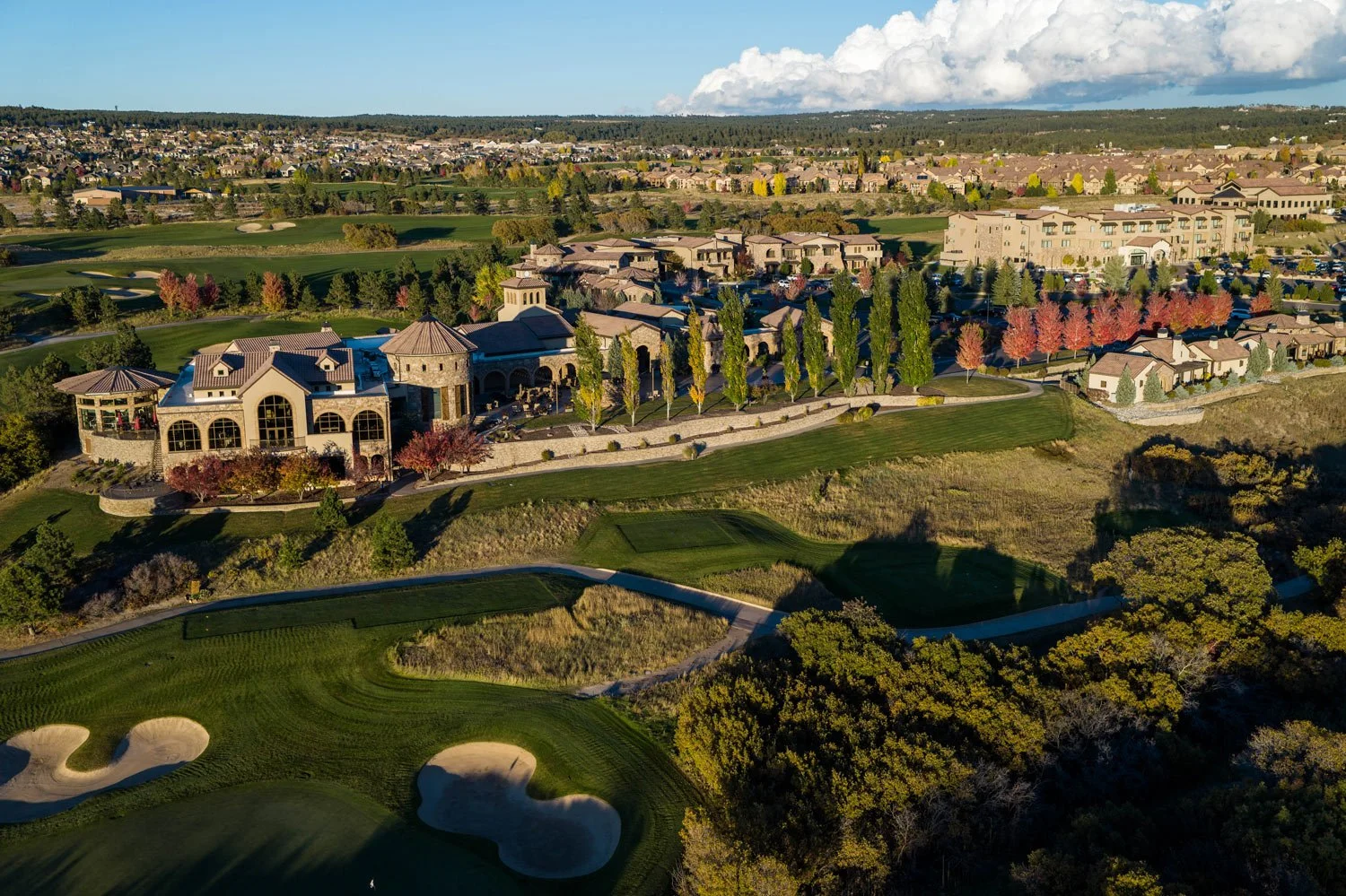 Aerial view of a large golf course with clubhouse, surrounding trees, residential buildings, and a distant cityscape under a partly cloudy sky.