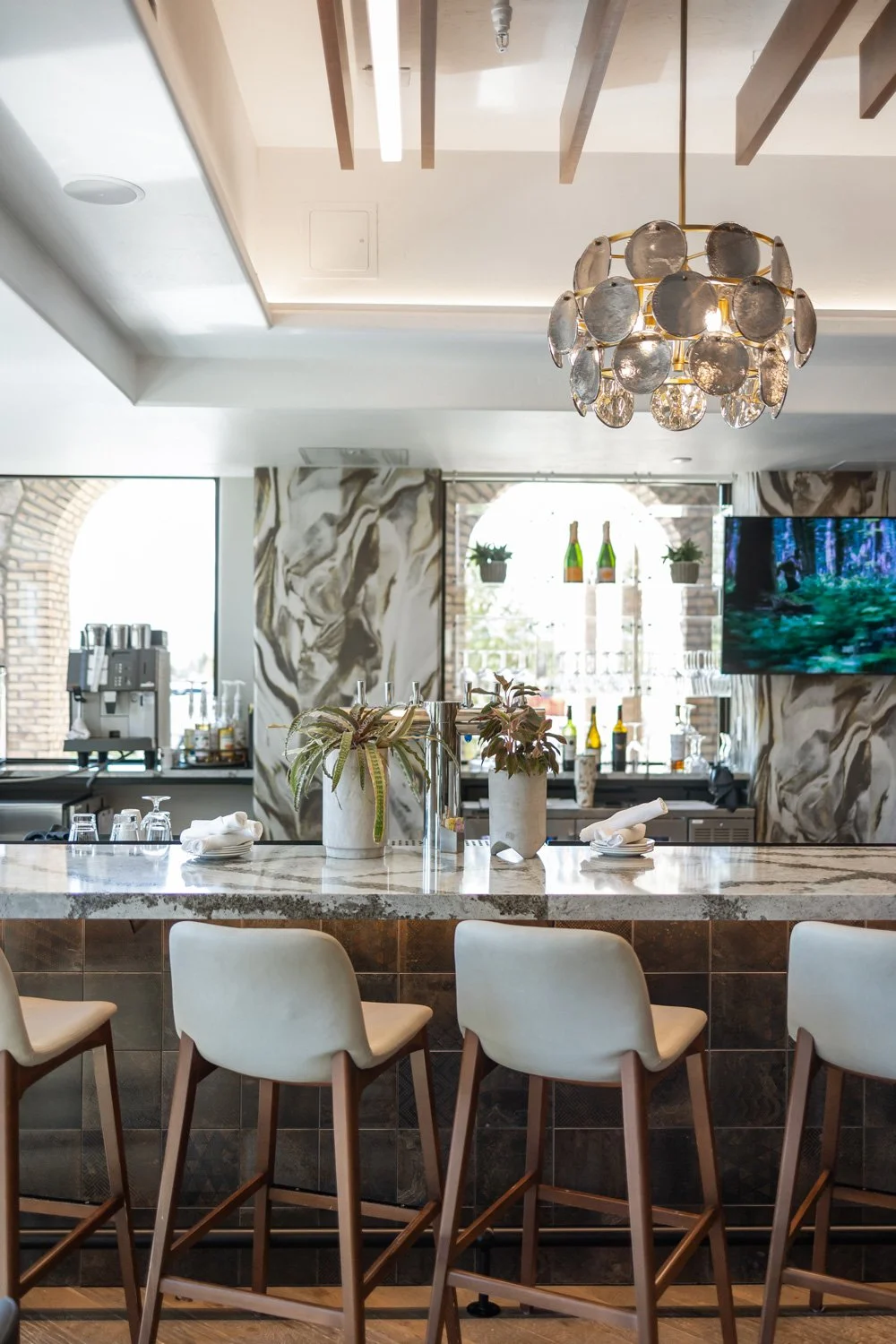 A modern bar or restaurant interior with a marble counter, bar stools, and decorative plants. There is a chandelier hanging from the ceiling, and a window with plants and bottles in the background.