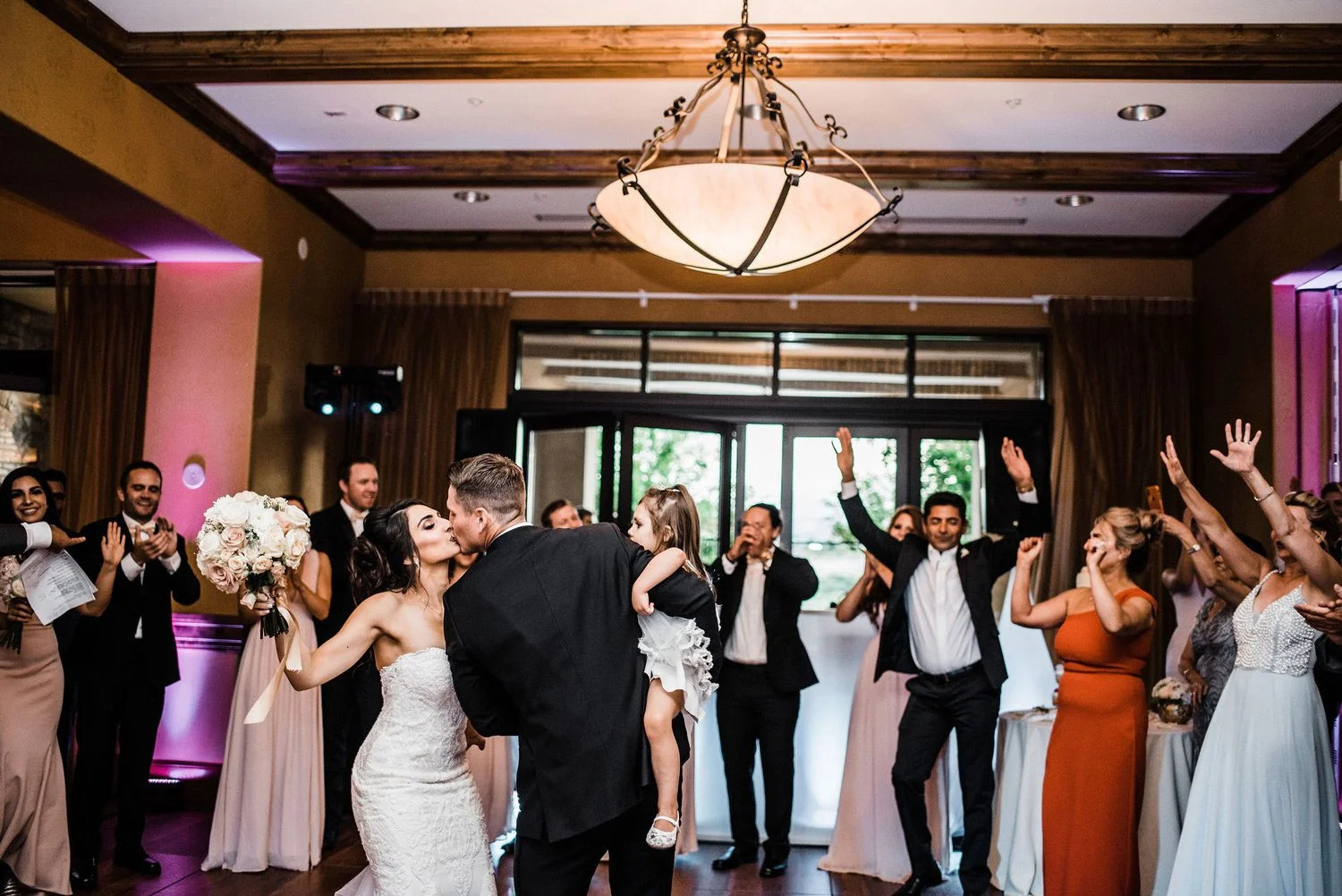 A wedding celebration with a bride and groom kissing in the center, surrounded by guests clapping and cheering inside a decorated venue with a chandelier and large windows.