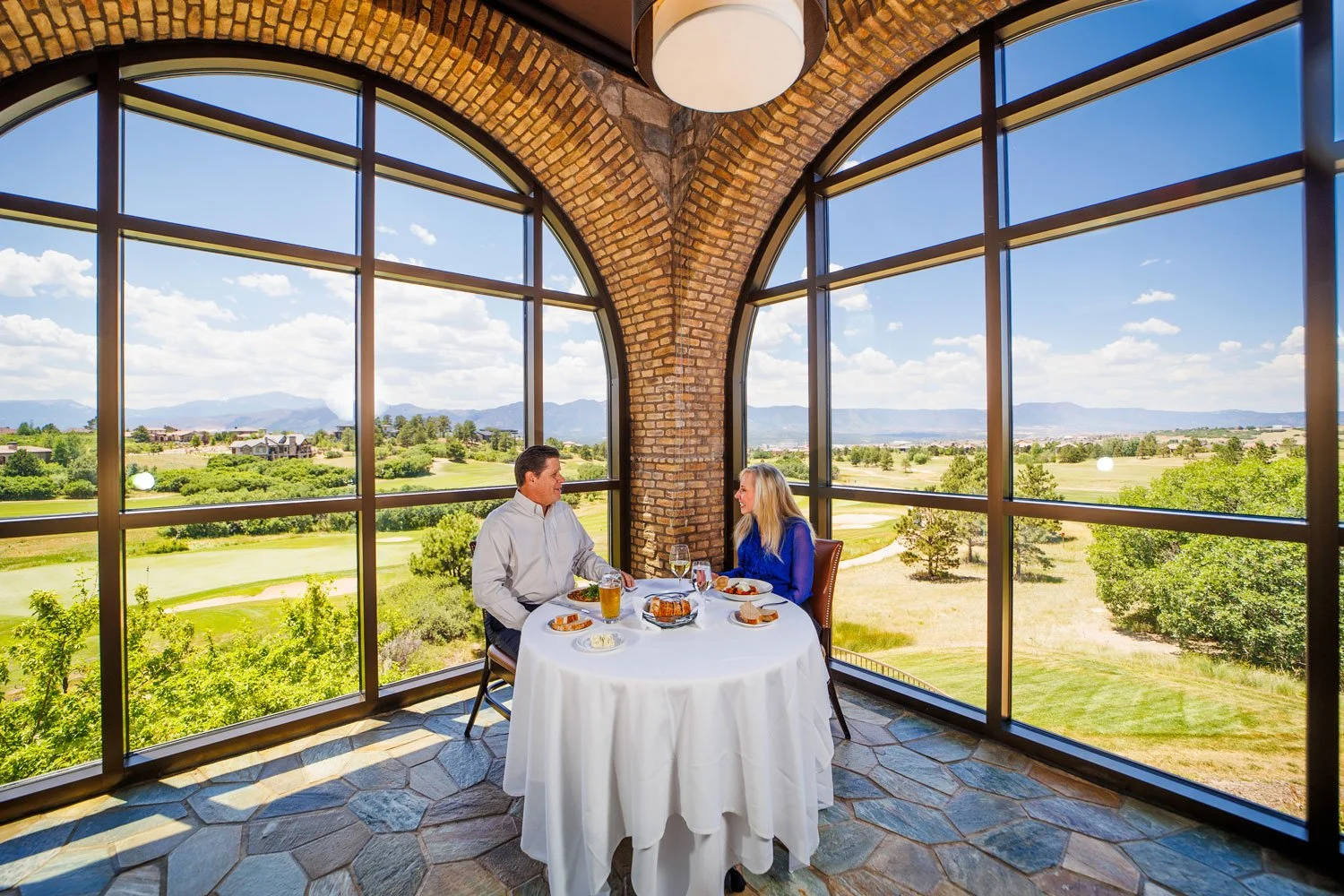 A man and a woman sitting at a round table with a white tablecloth inside a glass-walled room overlooking a scenic outdoor view of green fields, trees, and mountains on a sunny day.