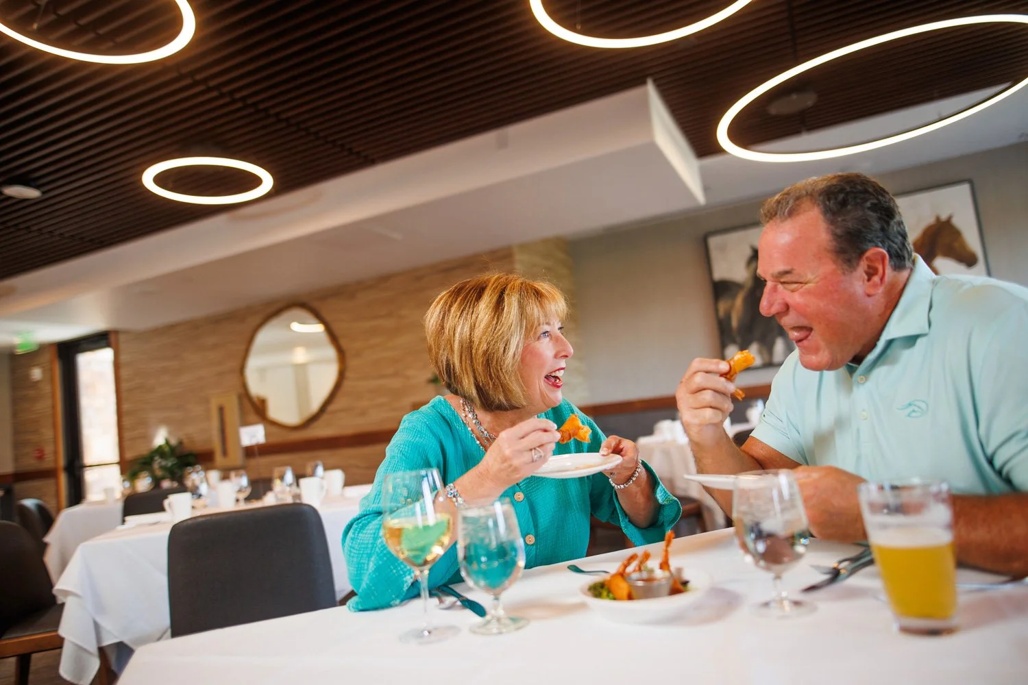A smiling older woman and a smiling older man enjoying a meal together at a restaurant. They are holding food and appear to be having a lively conversation. The table has wine glasses, a beer, and plates of food. The restaurant has modern decor with 