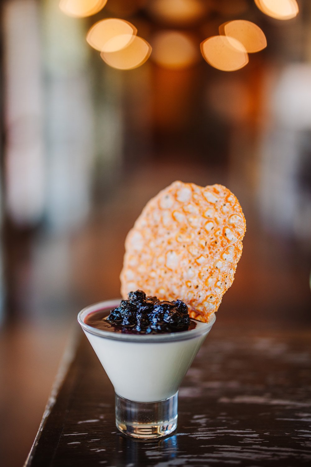 A dessert served in a glass cup with white pudding, topped with dark berry syrup and topped with a round rice cake and a rice cracker.