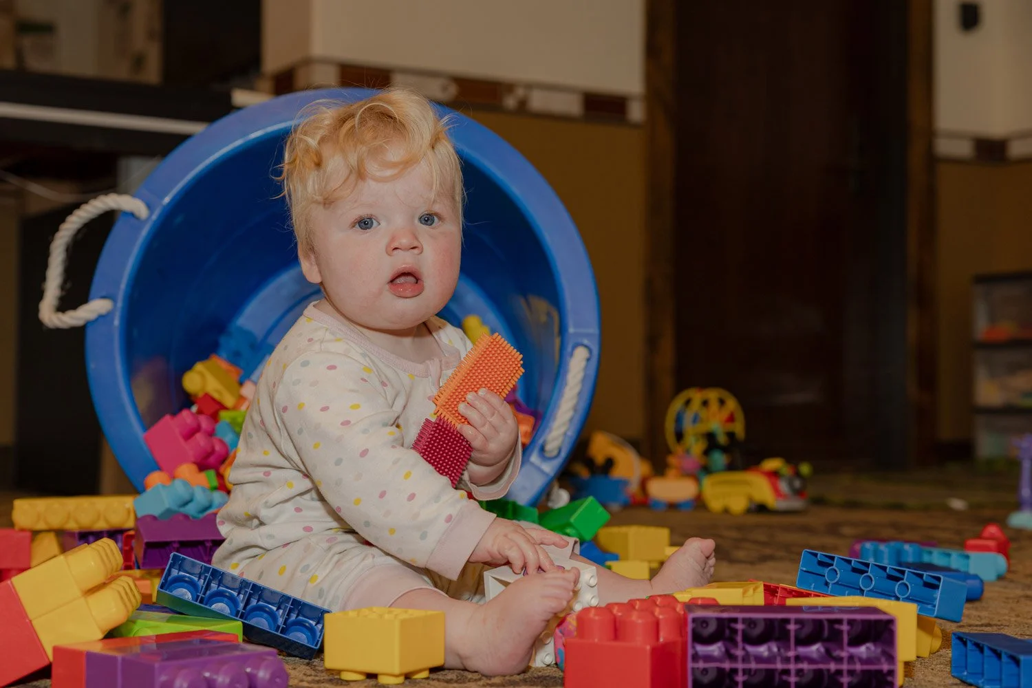A young child with blonde curly hair and blue eyes sitting on the floor amidst colorful plastic building blocks and toys, with a large blue bucket tipped over behind them.