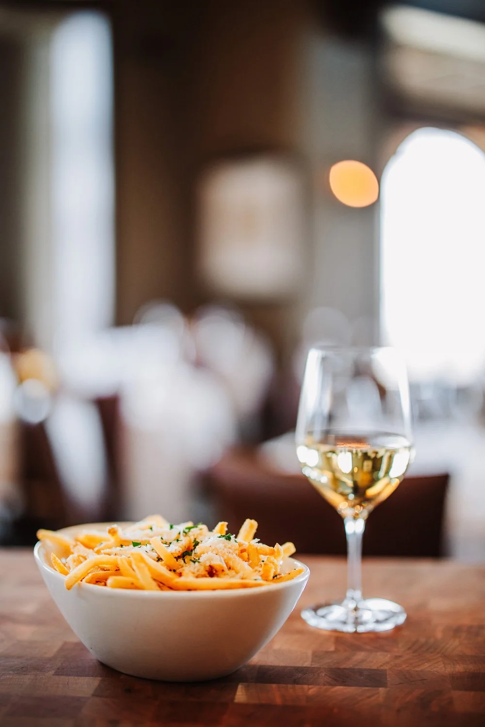 A bowl of fries with cheese and herbs next to a glass of white wine on a wooden table at The Steakhouse.