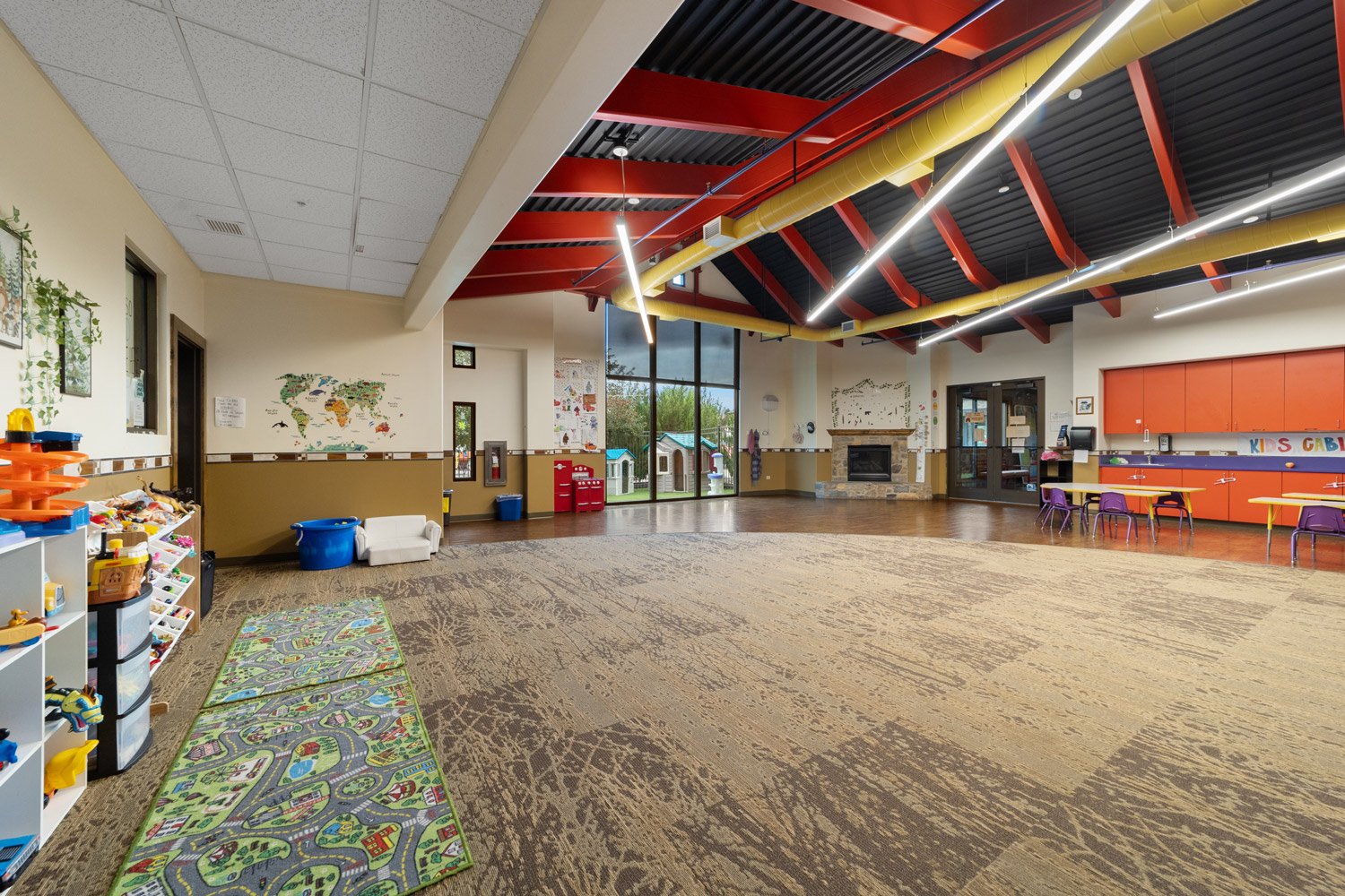 Large indoor activity room with colorful carpet, toy shelves, playhouse, and tables for children, featuring a high ceiling with exposed beams and ductwork, and large glass doors leading outside.