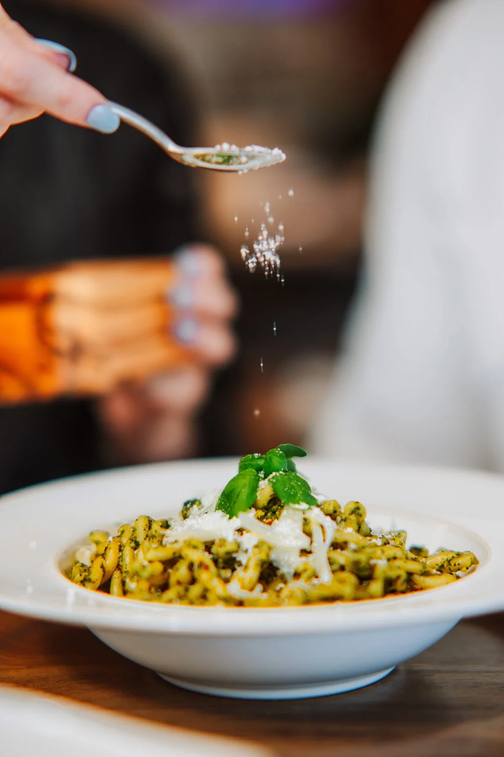 Close-up of a person sprinkling grated cheese on a bowl of pasta garnished with basil leaves.