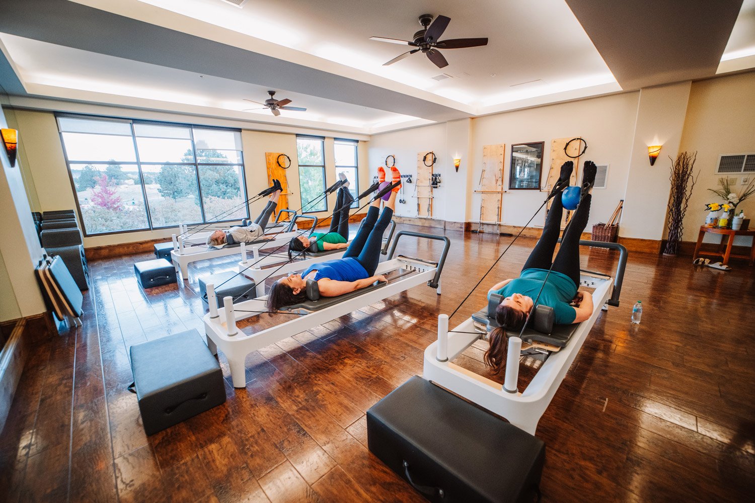 Women exercising on reformer Pilates machines in a fitness studio with large windows, wooden floors, and ceiling fans.