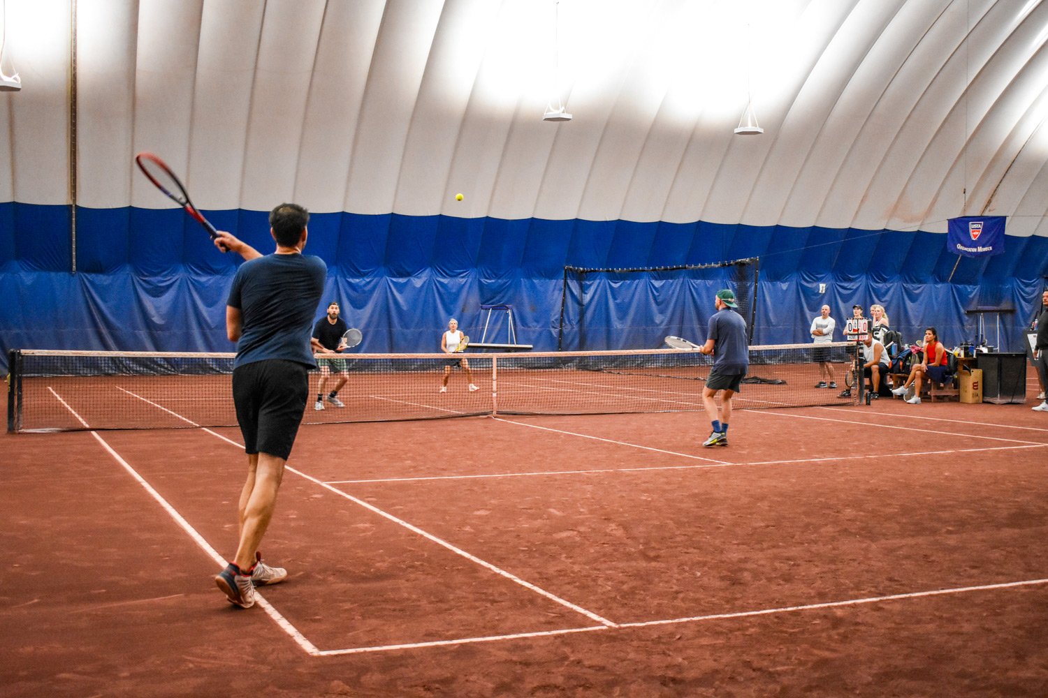 People playing doubles tennis on an indoor clay court with others watching on the side.