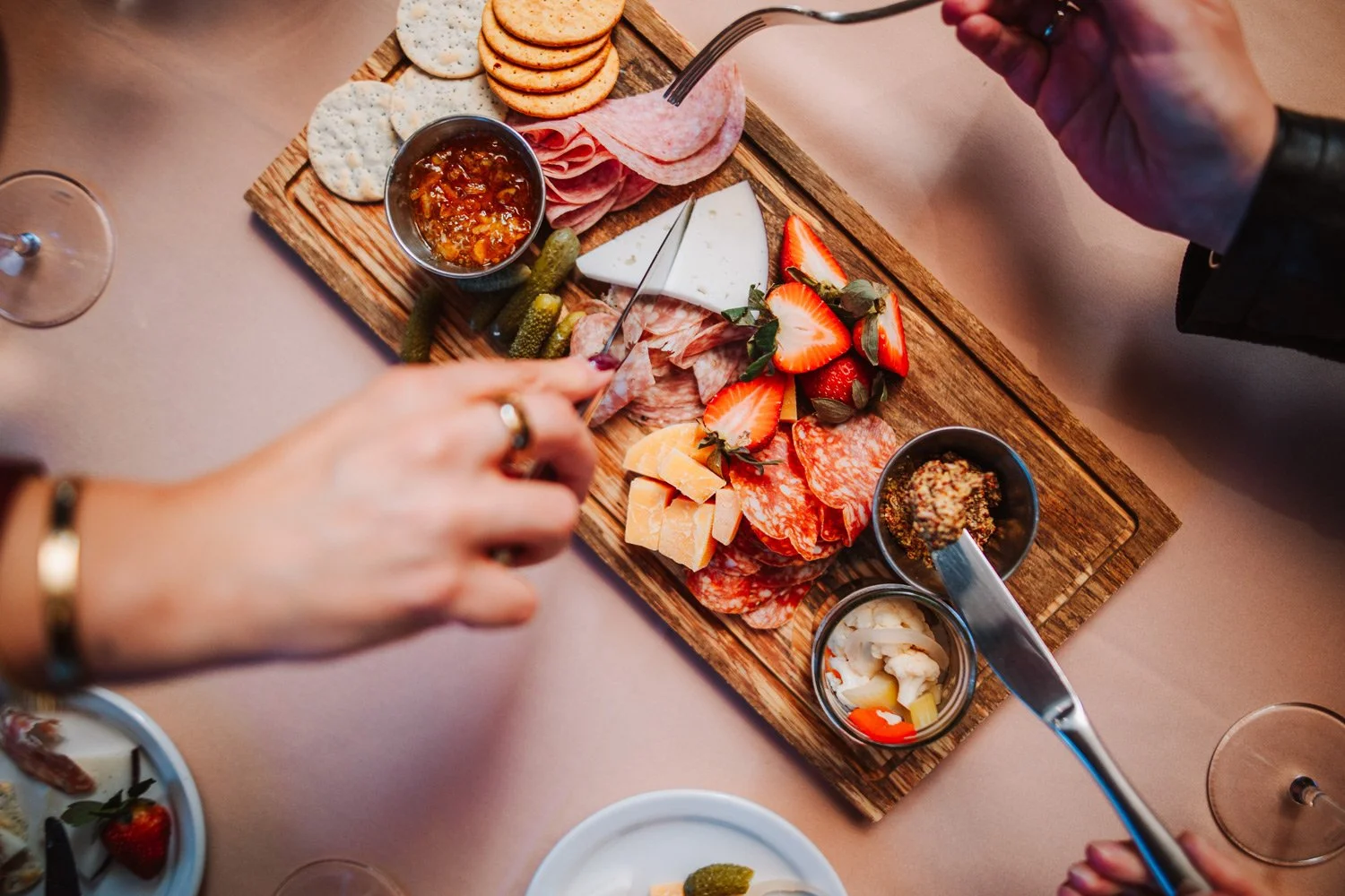A wooden serving board with cheese, strawberries, cured meats, crackers, and small bowls of jams and spreads, presented on a pink tablecloth.