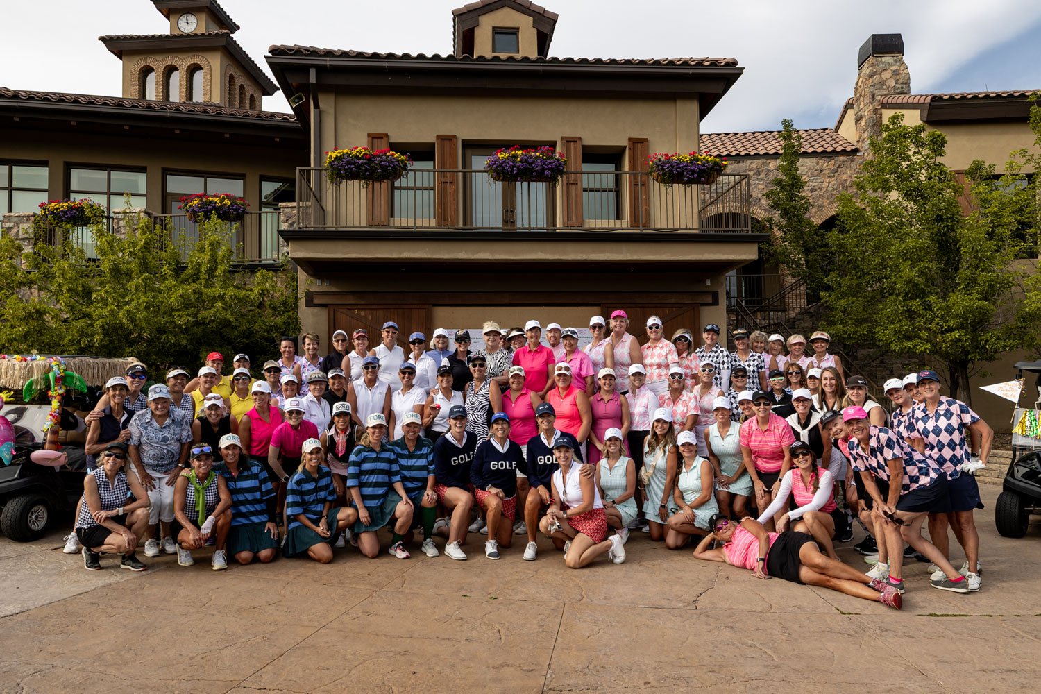 A large group of women, mostly wearing golf attire and caps, posing together for a photo outside a building with a stone and stucco exterior, balcony with flower boxes, and a clock tower in the background. Some women are kneeling in front, others are