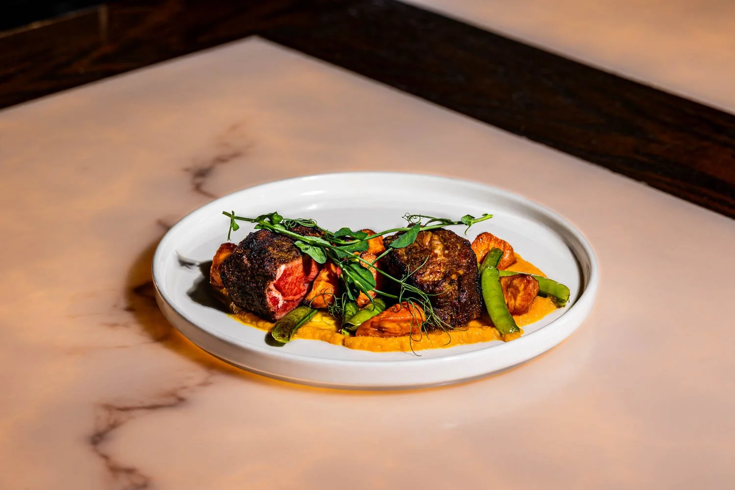 A white round plate containing a cooked beef tenderloin with a crispy exterior, sliced to show the pink interior, served on a bed of orange mashed sweet potatoes, garnished with fresh herbs and green beans, on a marble countertop.